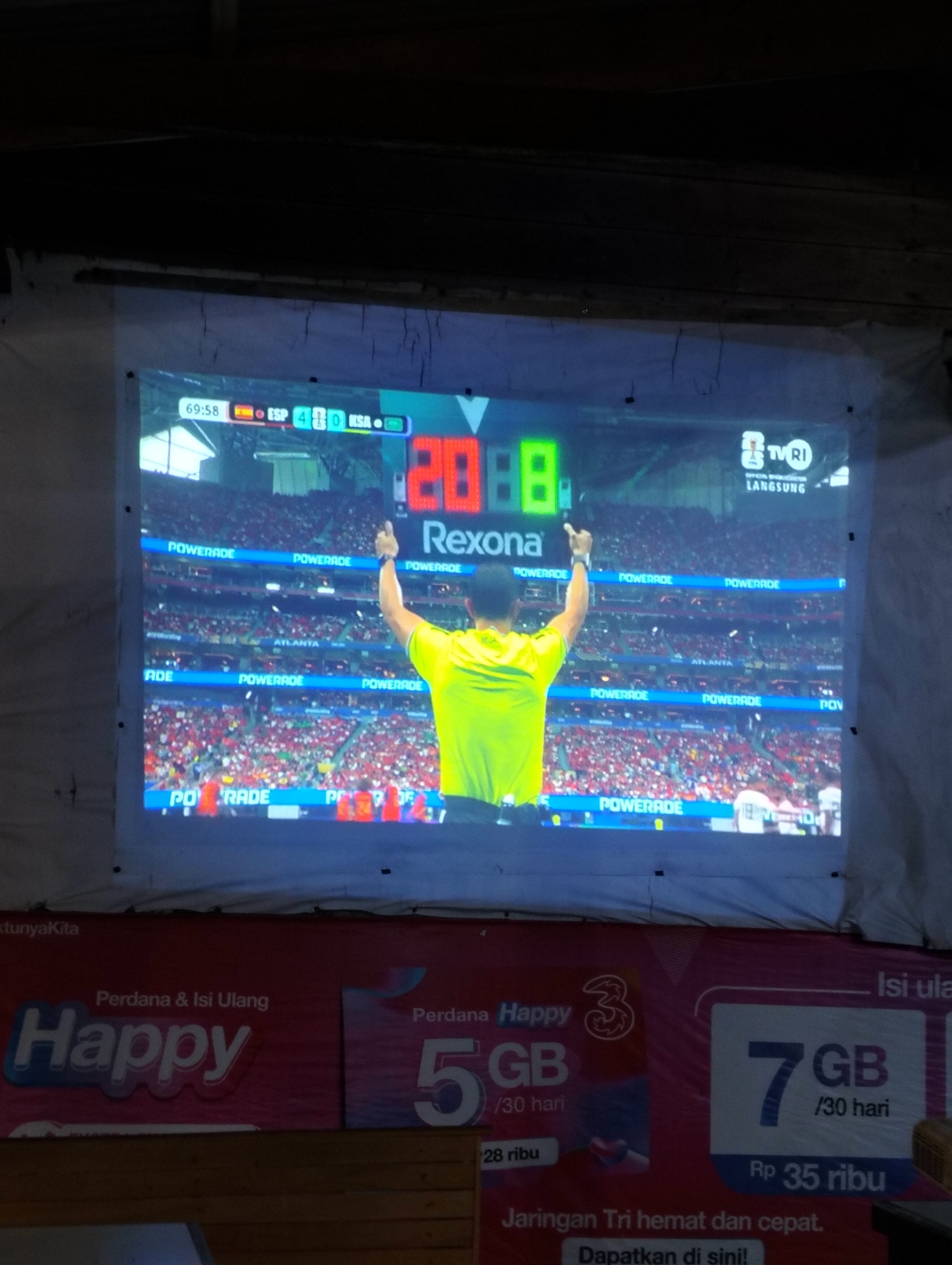A projected image displays a soccer match with a score of 4-0. A referee in a yellow jersey is visible, his arm raised. The screen shows advertisements for Powerade and a TV channel called TVRI. Below the projected image, there are advertisements for mobile phone data plans, including "Happy" and "7 GB / 30 hari" for Rp 35,000. The setting appears to be an indoor space, possibly a community hall or event space, with wooden planks visible at the bottom. The overall scene suggests a public viewing of a sporting event. The location is identified as Payagaboh, Indonesia.