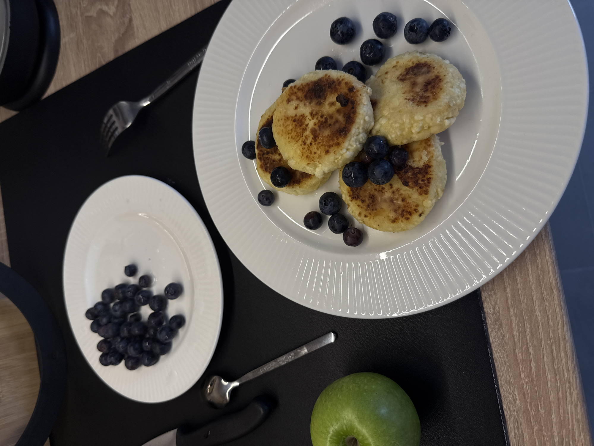 This is a top-down, slightly angled medium shot of a breakfast spread on a wooden table. In the foreground and center, a white circular plate with a fluted edge holds four golden-brown cheese pancakes, each topped with fresh blueberries. The pancakes are slightly irregular in shape, suggesting they are homemade. Several loose blueberries are scattered around the pancakes on the plate. To the left of this plate, partially in the frame, is a vibrant green apple. Further left and slightly behind the apple is a black handled knife, and next to it, a small, tarnished silver spoon. Above and to the left of the apple is another white circular plate, this one holding a pile of fresh blueberries. A silver fork lies on the table to the right of the main plate. The background is blurred, but appears to show a dark placemat and a glimpse of a dark appliance. There are no people present in the image. The scene depicts a simple, fresh breakfast setup. The image does not provide any information that would allow for the determination of its location in Moscow, Russia.