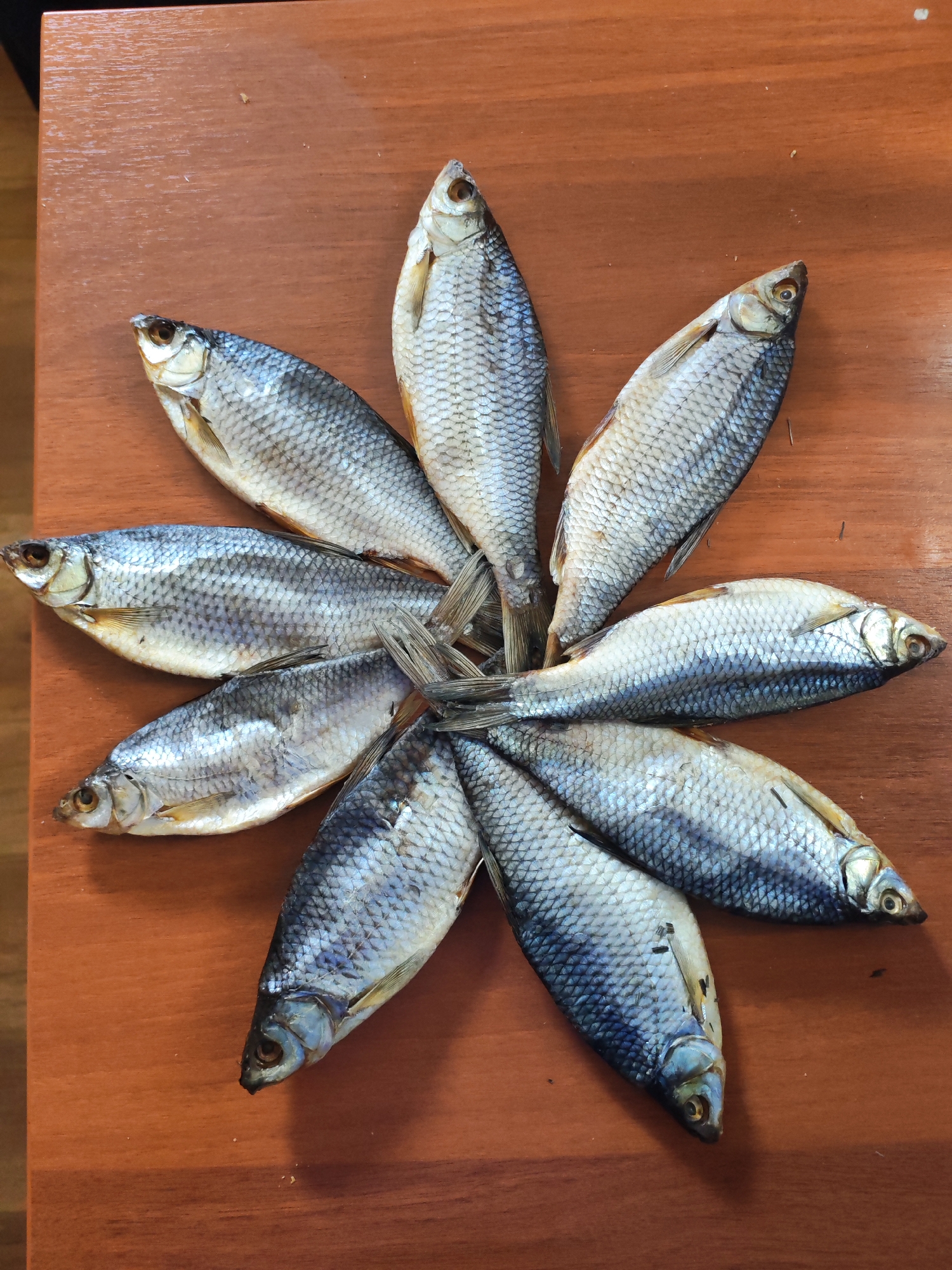 The image shows eight dried fish arranged in a circular pattern on a brown wooden surface. The fish are small and silvery, with visible scales and fins. The arrangement is centered on the surface, with the tails of the fish meeting in the middle. The background is a plain wooden surface with no other objects or distractions visible. The location of the image is Unknown, Unknown.