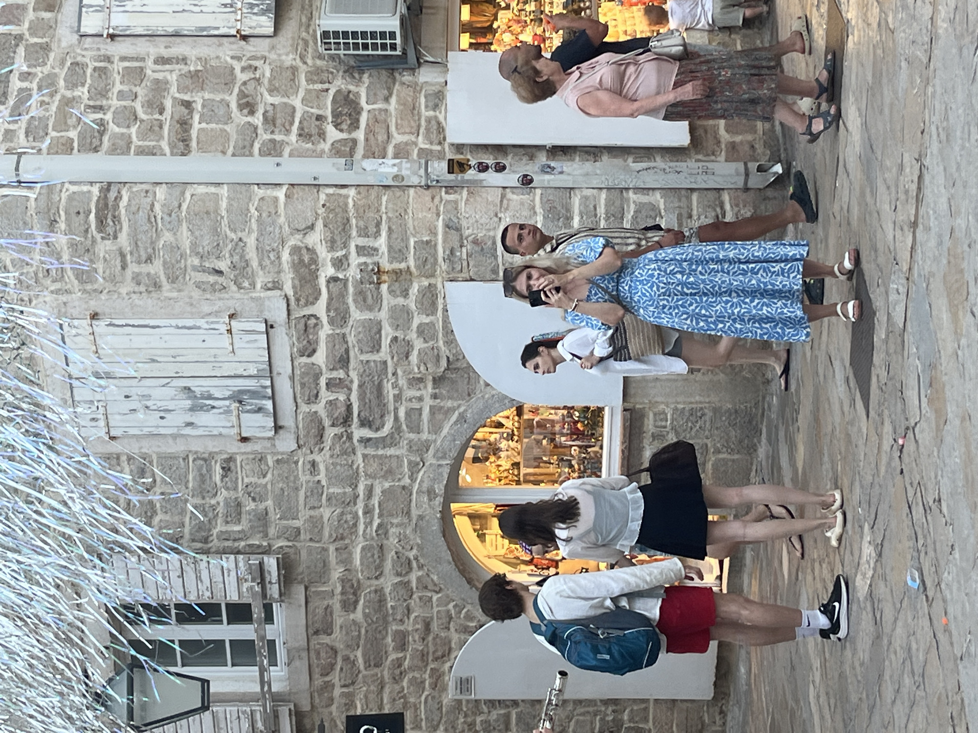 In Budva, Montenegro, on a cobblestone street, a group of people are gathered outside a stone building. The building has weathered wooden shutters on its windows and a visible air conditioning unit. Festive, shiny, iridescent decorations hang from above. In the foreground, a young man with a blue backpack and red shorts stands on the left, looking toward a shop window. Next to him, a young woman in a white top and black skirt holds a large dark bag. To her right, another young woman in a blue and white patterned dress is holding a smartphone up, as if taking a picture. She wears white sandals. Behind her, a man in a striped shirt looks forward, and further to the right, a woman in a pink top and patterned skirt walks away from the camera, carrying a white purse. In the background, a child in a light-colored shirt and shorts is visible near another shop entrance. The overall scene captures a moment of leisure and tourism in a Mediterranean setting.