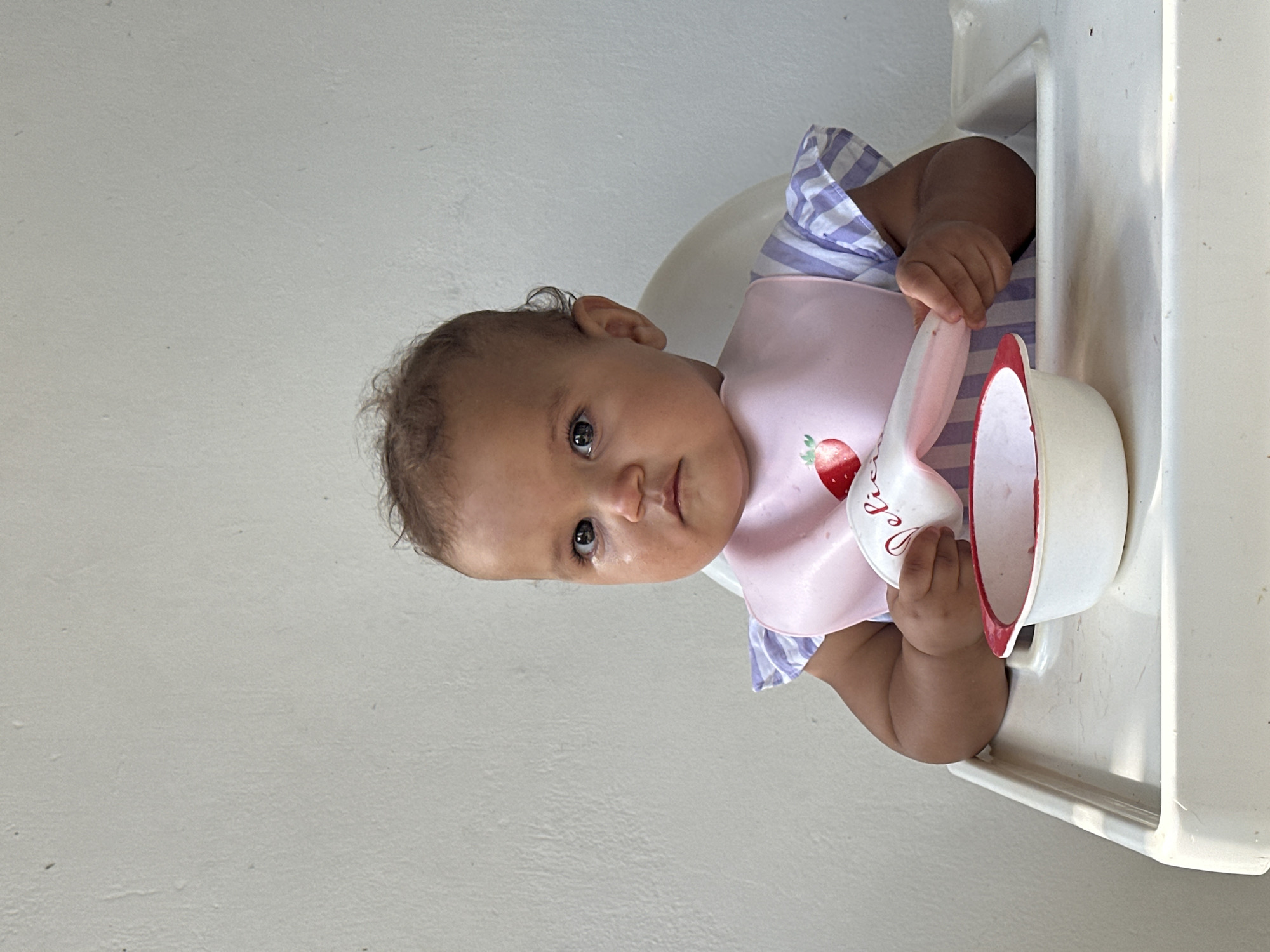 A baby is sitting in a high chair in front of a white wall. The baby is wearing a pink bib with a strawberry on it and a purple and white striped shirt. The baby has a spoon in its hand and a bowl in front of it. The baby is looking at the camera with a serious expression. The high chair is white and made of plastic. The bowl is white with a red rim. The background is a plain white wall.