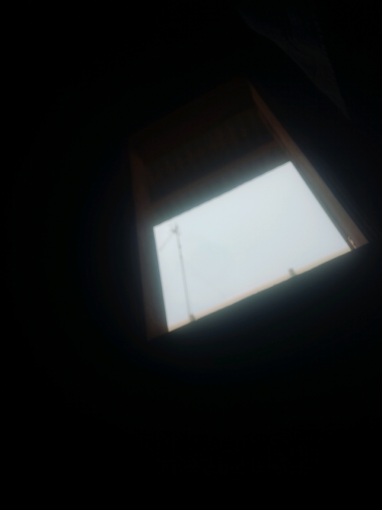 A dimly lit interior shot shows a window with a view of a light gray, overcast sky and a distant transmission tower. The window is rectangular and appears to be a skylight, set within a wooden frame. The foreground is almost entirely black, obscuring the rest of the interior space. The image is grainy, and the focus is soft, suggesting it was taken in low light conditions. The location is not identifiable from the visual information.