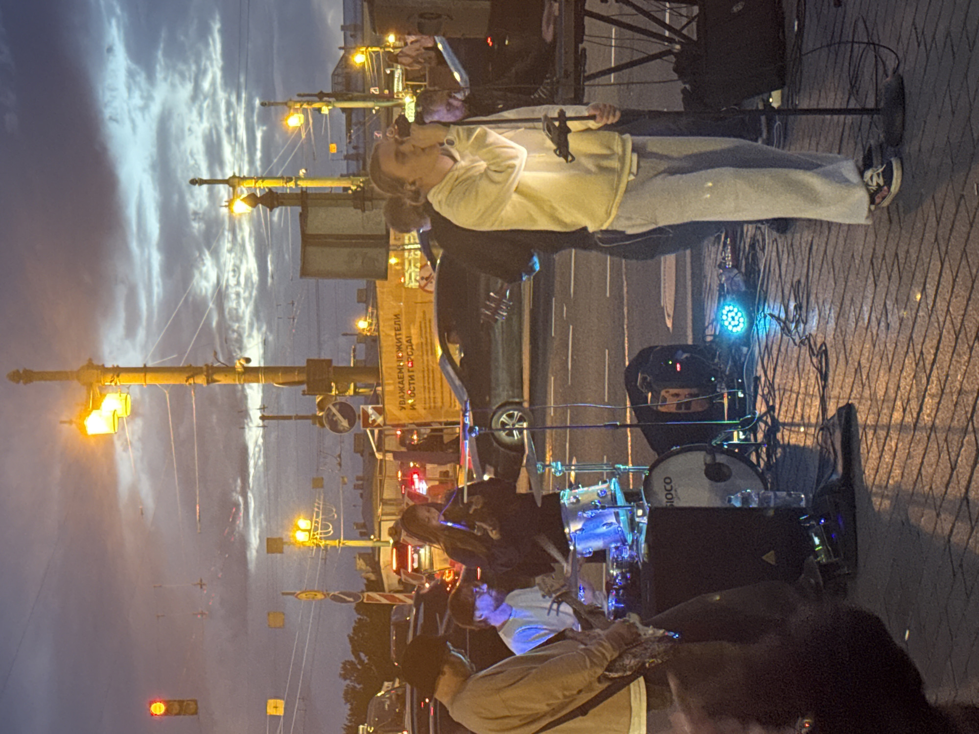 The image captures a street performance in Saint Petersburg, Russia, likely during twilight hours. A band consisting of at least three individuals is performing on a paved city street, with cars and traffic lights visible in the background. The central figure is a young person singing into a microphone, dressed in a light yellow hoodie and grey sweatpants. To their left, a person wearing a dark hoodie and holding a phone is playing a drum kit. Another person, seen from the back and wearing a dark beanie and light brown hoodie, is playing a guitar. In the background, street lamps with warm yellow light illuminate the scene, and a traffic light shows a red signal. The sky is cloudy, with hues of blue and grey. A yellow sign with Cyrillic text is partially visible on the right side of the frame. The overall atmosphere suggests a spontaneous or planned outdoor musical event in an urban setting.