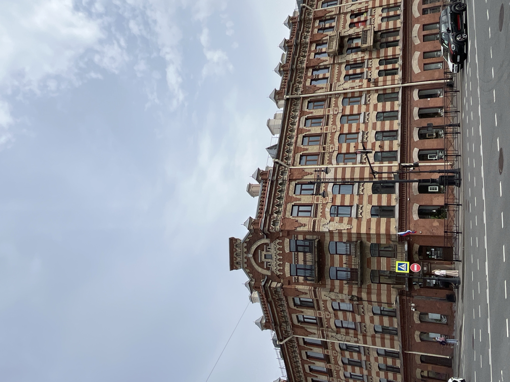 The image features a tall, ornate building in Saint Petersburg, Russia, composed of alternating red and beige horizontal stripes. The building has multiple windows on each floor. A black car is parked along the edge of the street next to the building. A road with white lane markings runs vertically on the right side of the image. A pedestrian crossing sign and a "no entry" sign are visible near the building entrance, which is decorated with a Russian flag. Several people stand near the entrance. The sky is overcast.