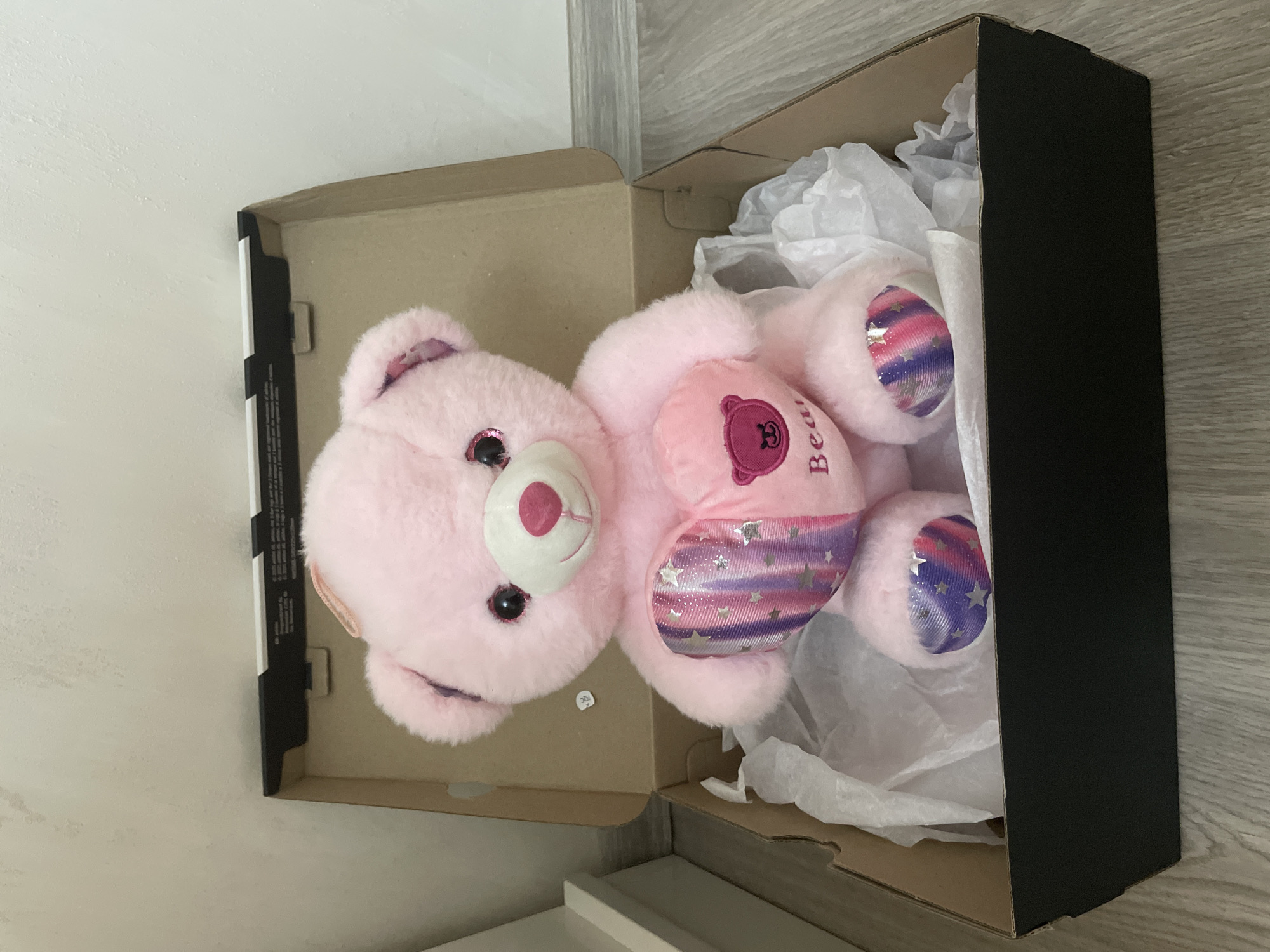 A pink, fluffy teddy bear is placed inside an open cardboard box. The teddy bear features large, dark, glittery eyes and a pink nose. On its belly, there is a heart shape containing a letter (appears to be 'B' or 'R') and the word "Bear" written vertically. Its paws are made of a shiny, iridescent material with pink, purple, and blue stripes, decorated with small silver stars.
The teddy bear rests on crinkled white tissue paper, which fills part of the box. The box has a brown interior and a black exterior panel on one side. The background shows a light-colored wall and a light-toned wooden or laminate floor, indicating an indoor setting. The arrangement suggests the teddy bear is either being prepared as a gift or stored. Some indistinct white markings, possibly text or a barcode, are visible on the black interior flap of the box, but are not legible.
