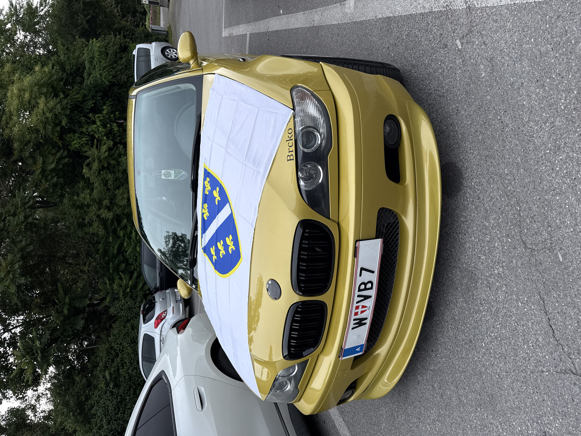 A gold BMW E46 sedan is parked on an asphalt surface. A white flag with a blue fleur-de-lis crest is draped over the hood. The car's license plate reads "W VV B 7". The word "Brcko" is visible on the right headlight. In the background, trees and other vehicles are present, including a white sedan and a silver hatchback. The overall environment suggests an outdoor parking area, possibly in a suburban or commercial district.