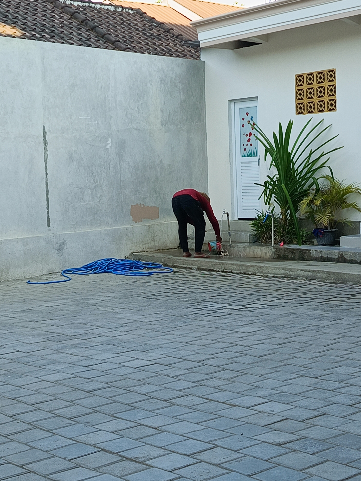 A person in a red shirt and black pants is watering plants with a blue hose. The person is bending down and holding a red and white watering can under a faucet. The scene takes place outdoors on a paved area in front of a white building with a door decorated with floral imagery. A decorative yellow vent is visible on the wall of the building. A large blue hose is coiled on the ground to the left of the person. The building has a tiled roof. The time of day appears to be daytime, with natural light illuminating the scene. The location cues suggest a tropical climate, consistent with Kuta, Indonesia.