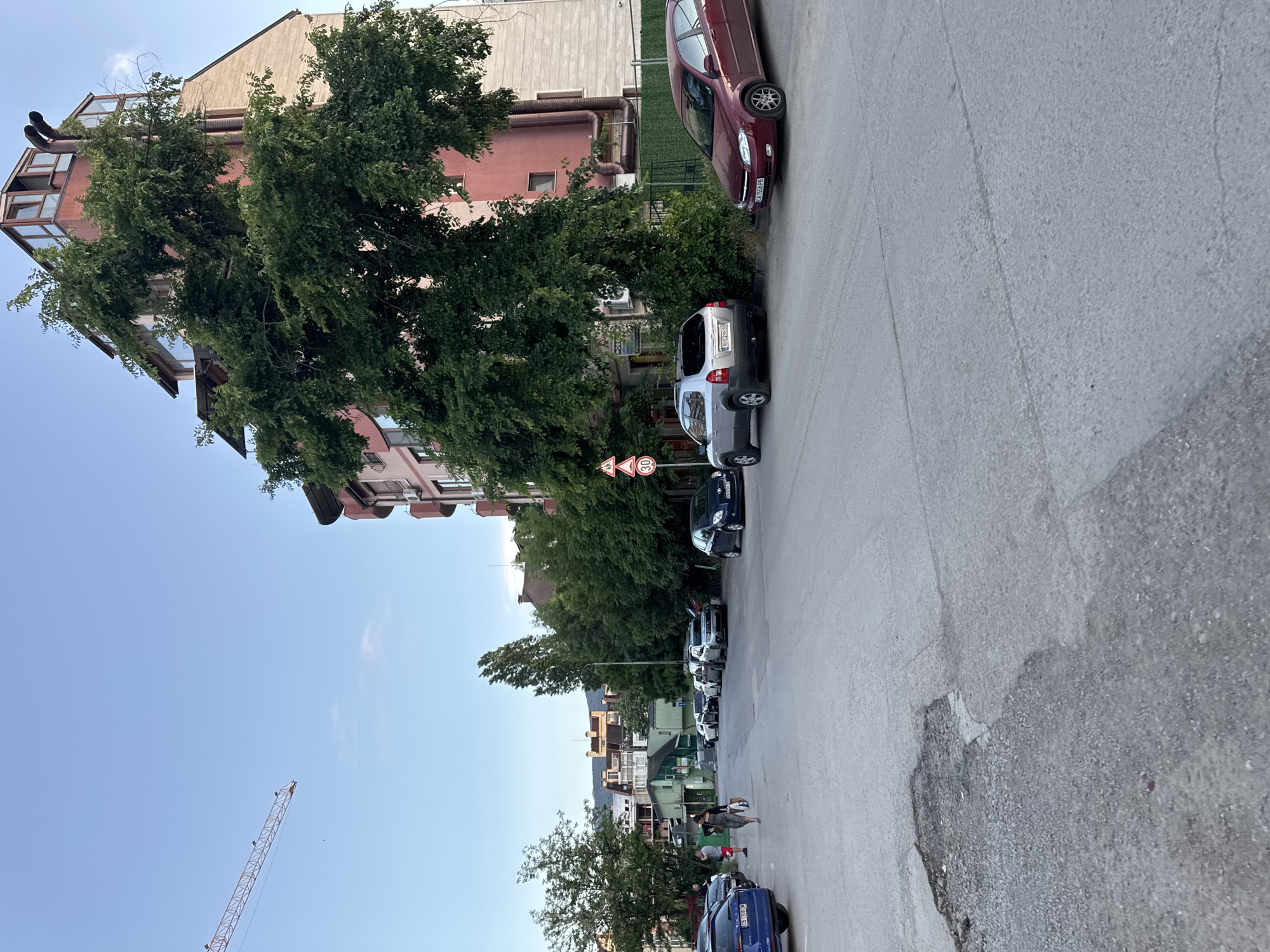 The image depicts a street scene in Sofia, Bulgaria, showing a residential area with buildings, trees, parked cars, and a blue sky. A red brick building with angled, modern architectural extensions is partially visible on the left. A line of trees obscures some of the buildings and lines the side of the road. Cars of various colors are parked along the right side of the road. The road itself appears to be made of asphalt. A 30 km/h speed limit sign is visible. In the background, more buildings are visible, and a crane is present in the distance. Two people are walking on the sidewalk.