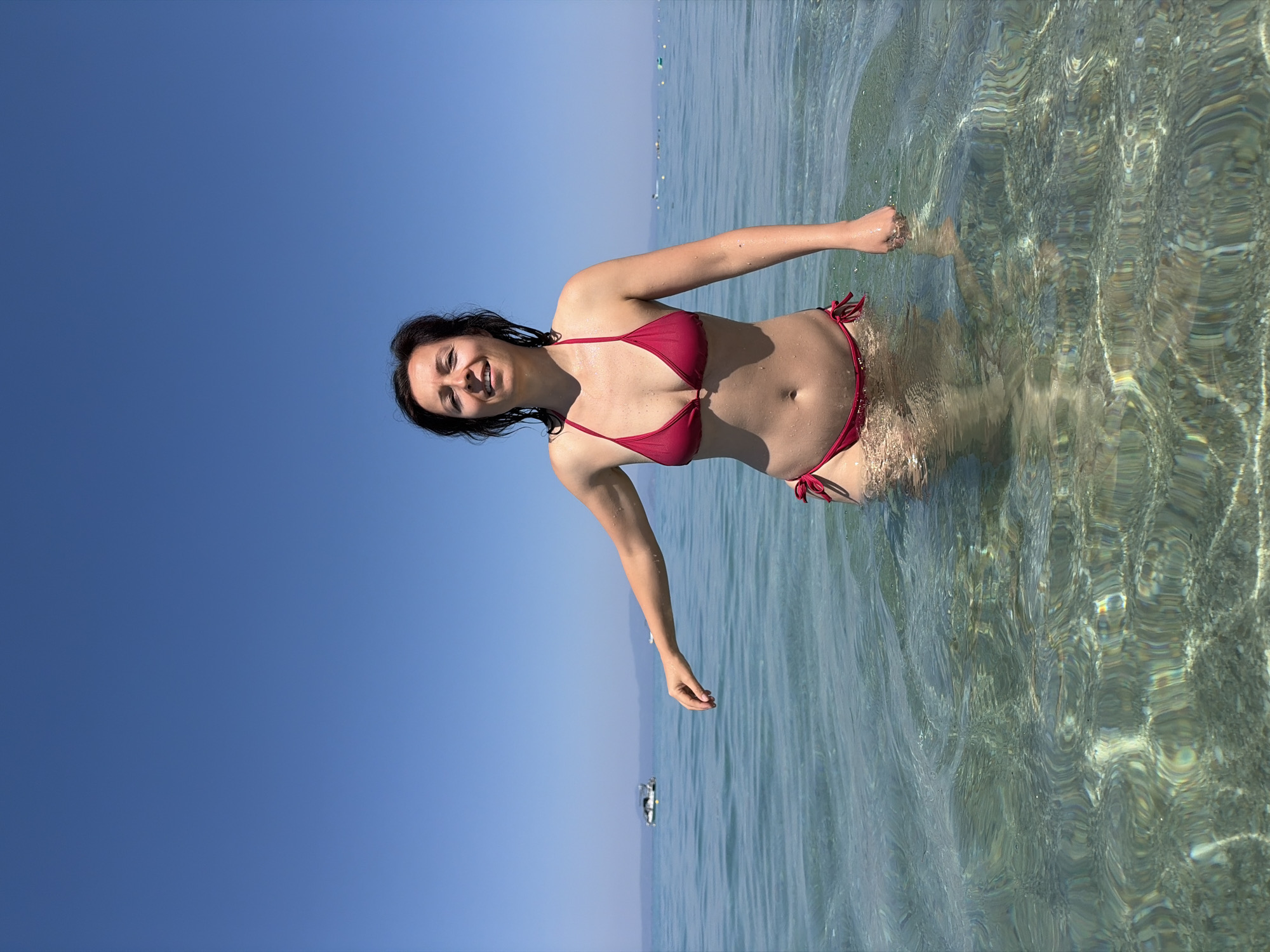 A woman in a red bikini stands in shallow, clear water under a bright blue sky. Her body is partially submerged, and her arms are extended outwards. The water's surface is rippled, creating distorted patterns of light and shadow on the sandy seabed below. In the distance, the horizon is visible, with faint outlines of land and a few small boats. The scene appears to be set in a coastal area, likely a beach or a bay. The sunlight suggests it is daytime, and the clear sky indicates fair weather. The woman's expression is relaxed and happy, with a slight smile on her face. There is no visible text in the image. The context provided indicates the location as Pallini Municipal Unit, Greece, suggesting a Mediterranean setting.