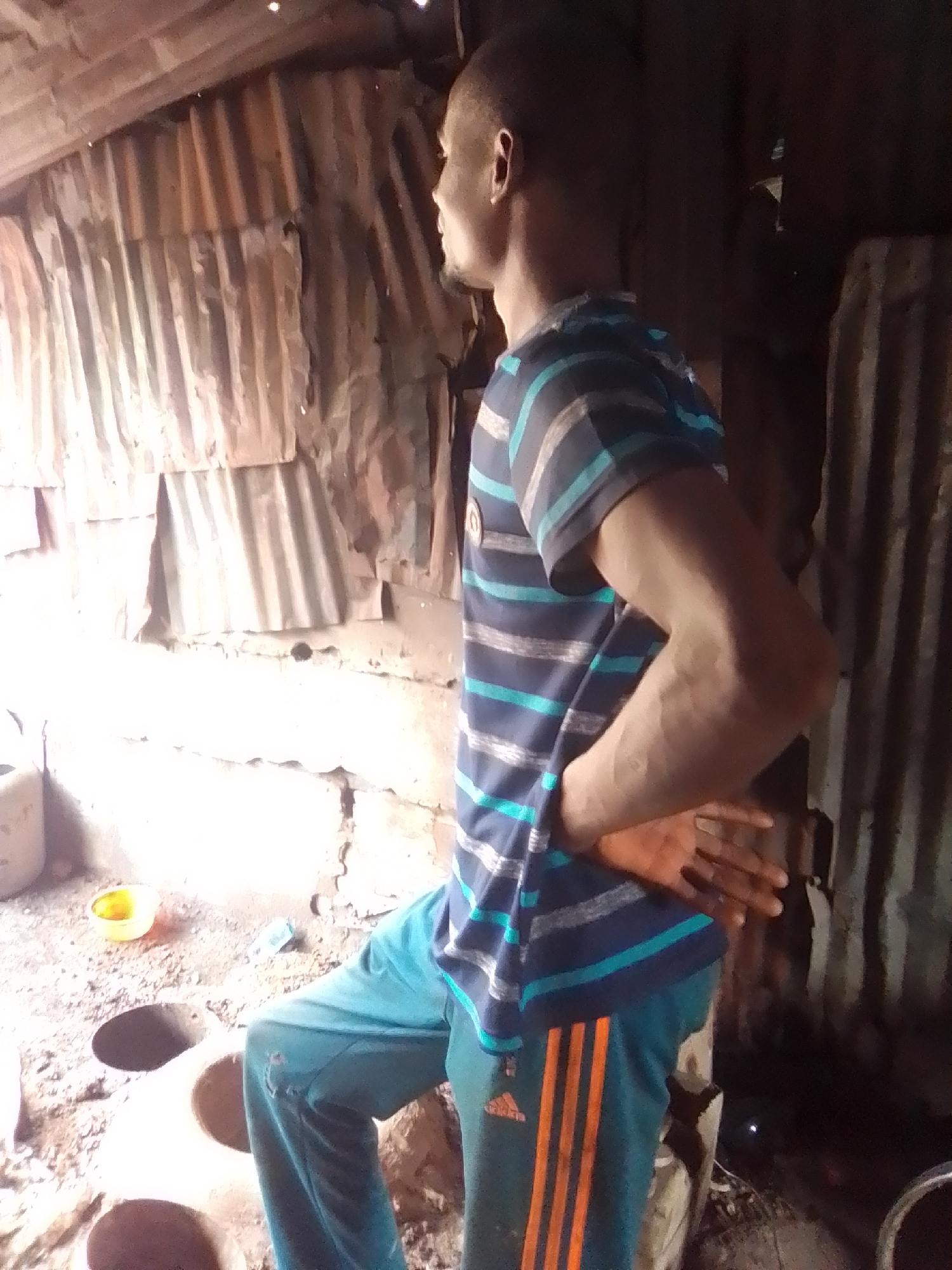 A person stands in what appears to be a makeshift shelter constructed with corrugated metal sheeting and wood. The interior is dimly lit, with a bright light source to the left of the frame. The floor is dirt and scattered with debris. The person is wearing a striped t-shirt and turquoise sweatpants with orange stripes. They are looking off to the right, with one hand resting on their hip. In the foreground, there are what appear to be clay ovens or kilns in the dirt floor. A small yellow bowl is visible on the ground to the left of the ovens. The overall impression is of a rural or informal settlement environment.