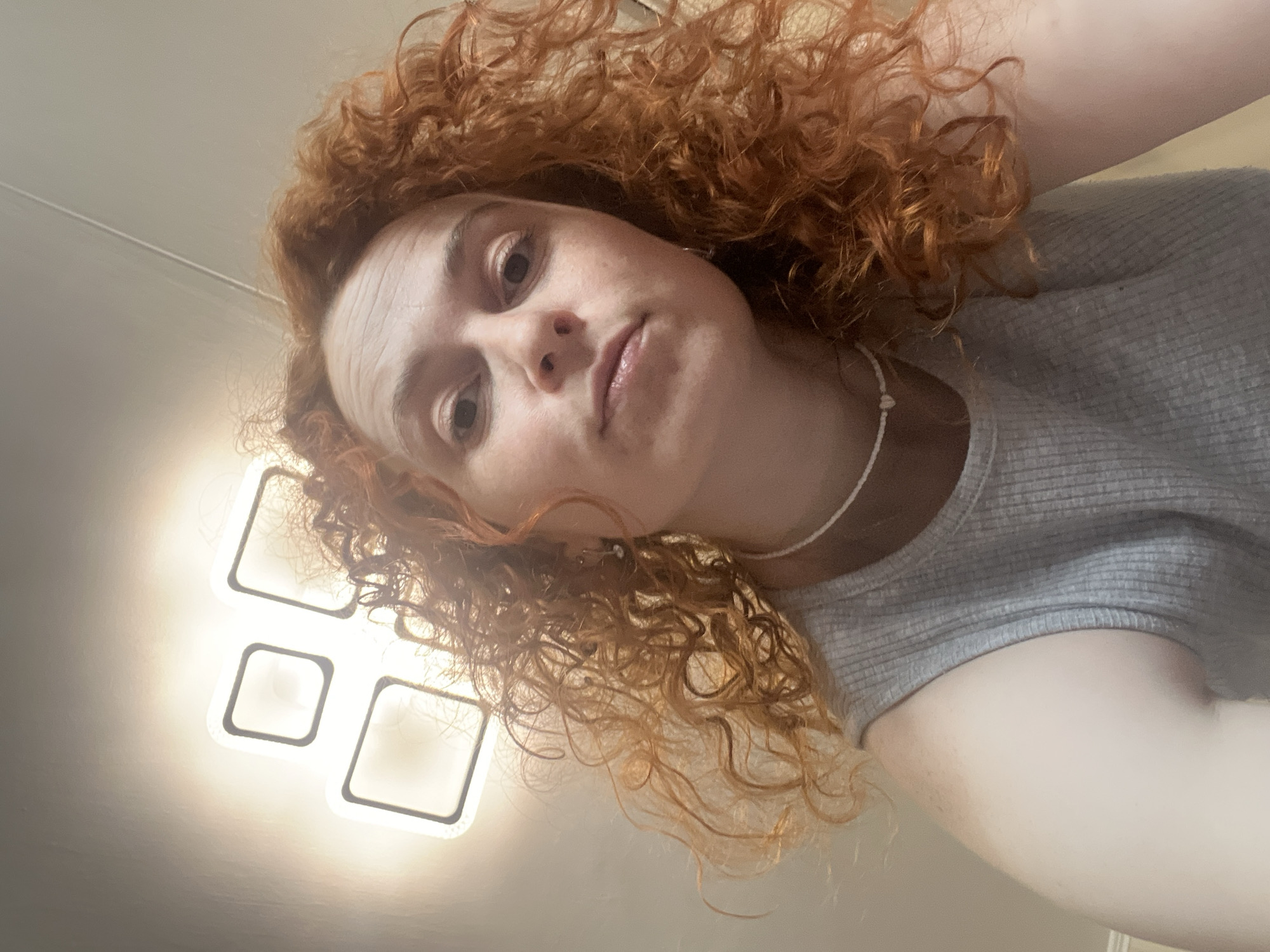 A young woman is captured in a close-up, head-and-shoulders selfie, with her head tilted to the right. She has voluminous, curly reddish-brown hair that frames her face. Her expression is neutral, with her lips slightly parted and appearing glossy. She wears a light grey, ribbed short-sleeved top and a simple white beaded necklace.
The activity taking place is a self-portrait (selfie). The scene is indoors, likely a room in Chaikovsky, Russia. The background primarily shows a light-colored ceiling or wall. A distinctive modern light fixture, composed of three glowing square elements outlined in dark frames, is prominently visible on the ceiling behind and to the left of her head, casting a warm glow. The light from this fixture appears to be a primary source of illumination. There are no visible texts or specific location cues beyond the architectural details of the light fixture and the indoor setting. The time of day is not discernible, given the reliance on artificial lighting.