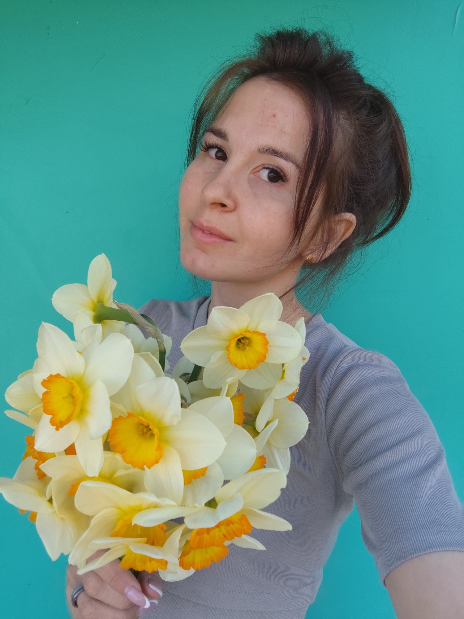 A young woman stands in front of a turquoise wall, holding a bouquet of white and yellow daffodils. Her hair is pulled up in a messy bun, with some strands falling around her face. She wears a gray long-sleeved shirt. She has light skin and brown eyes, and a subtle smile. She wears a ring on her left ring finger and her nails are painted with a French manicure style. The backdrop is a plain, textured turquoise wall. The scene is well-lit. The location is not determined to be Фоки, Russia.