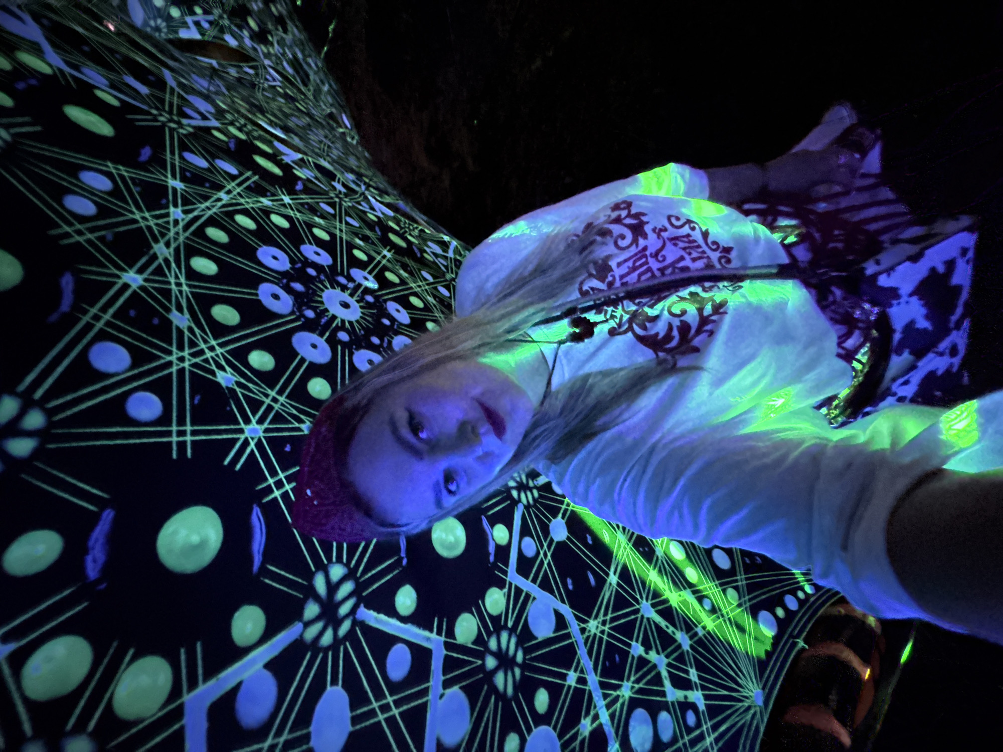 The image captures a selfie of a young woman with blonde hair, wearing a red bandana, a white t-shirt, and a patterned skirt. She is posing in front of a backdrop with a black surface adorned with intricate geometric patterns in green and blue, illuminated by what appears to be blacklight. The patterns consist of lines and circular shapes, creating a psychedelic effect. The lighting in the scene is predominantly blue and green, casting a vibrant glow on the woman and her clothing. The woman is looking towards the camera with a subtle smile. The background suggests an indoor environment, possibly a club or an art installation, given the nature of the lighting and decor. The location mentioned, Myl'niki, Russia, is not visually discernible from the image itself, as there are no identifiable landmarks or contextual clues within the photograph to confirm this specific place. The overall impression is one of a lively and visually stimulating atmosphere.