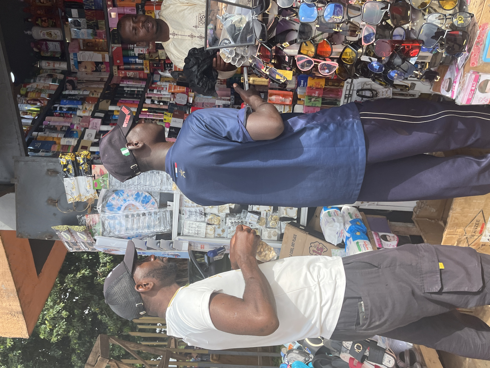 The image depicts a street market scene in Jos, Nigeria, with a focus on a stall selling a variety of goods, including cosmetics, sunglasses, and small accessories. Two men, dressed casually in t-shirts and trousers, are visible. One man, wearing a black baseball cap and a white t-shirt, stands on the left side of the frame and appears to be examining small items in his hand. The other man, wearing a navy blue t-shirt and a black baseball cap with a Nigerian flag emblem, is facing the stall. A third man, likely the vendor, is partially visible behind the stall, wearing traditional clothing and interacting with the customers. The stall itself is densely packed with shelves displaying numerous bottles of cosmetics and personal care products, as well as a prominent display of sunglasses. The environment suggests an outdoor urban setting, likely a busy commercial area within Jos.