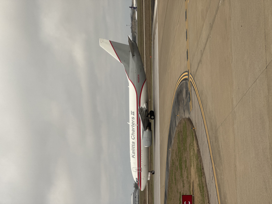 A white and grey Kalitta Charters II Boeing 737-type cargo aircraft, registered N332CK, is positioned on a concrete airport taxiway at Nashville International Airport (BNA) in Nashville, Tennessee, United States. The aircraft's fuselage is white with a prominent red stripe running horizontally, while its tail section is grey with a white top and a diagonal red stripe. Two turbofan engines are mounted under the wings. The foreground shows a curved section of concrete taxiway marked with double yellow lines, indicating taxiway edges, and white lines for runway hold positions. Adjacent to the taxiway on the left is a patch of dormant, brown-green grass with a red square sign displaying a partial white letter 'C'. The sky is uniformly overcast and grey. In the distant background, faint structures consistent with airport infrastructure and the purple tail of another aircraft are visible. No people are present in the scene.