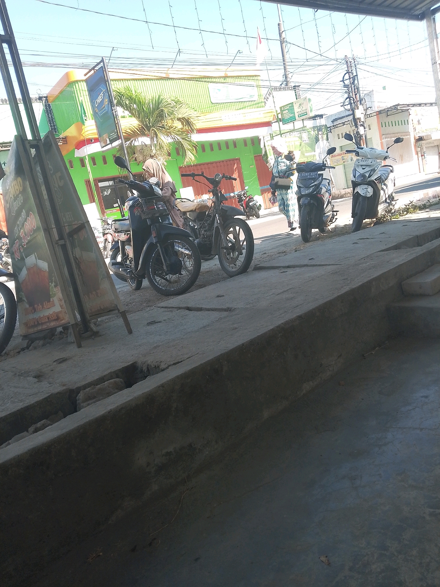 The image shows a street scene in Jampet, Indonesia, with several motorcycles parked along the sidewalk. A person wearing a brown headscarf and a light-colored shirt is standing next to a black motorcycle. Another person is walking behind them, wearing a patterned blue and white outfit. In the background, there is a green building with orange trim and a sign that reads "CHIEF". There are also utility poles and wires overhead, indicating an urban or semi-urban environment. A sign with an image of iced tea and text in Indonesian is visible on the left side of the image. The ground is paved with concrete, and there are some plants growing along the edge of the sidewalk. The overall impression is a snapshot of daily life in a bustling Indonesian town.