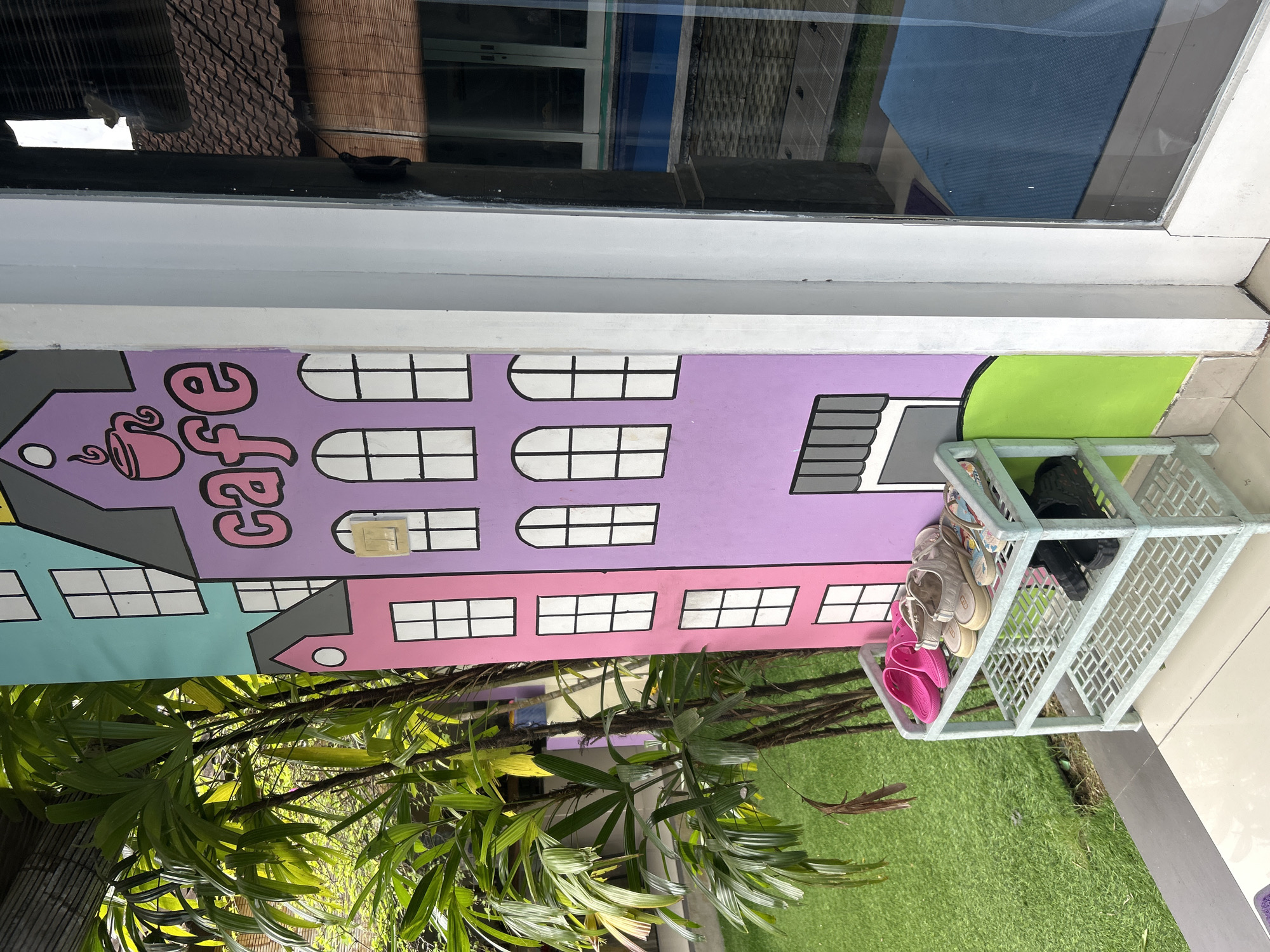 The image depicts an outdoor scene in Kuta, Indonesia, featuring a vibrant mural of colorful buildings painted on a wall. In front of the mural, a light blue plastic shoe rack holds several pairs of footwear, including pink flip-flops, beige sandals, and black slip-on shoes. To the left of the shoe rack, lush green artificial turf is visible, with palm fronds from a tree extending into the frame from the upper left. To the right, a glass door reflects the surrounding environment, showing a glimpse of a building with a tiled roof and bamboo blinds. The overall impression is a welcoming and somewhat whimsical entrance area.