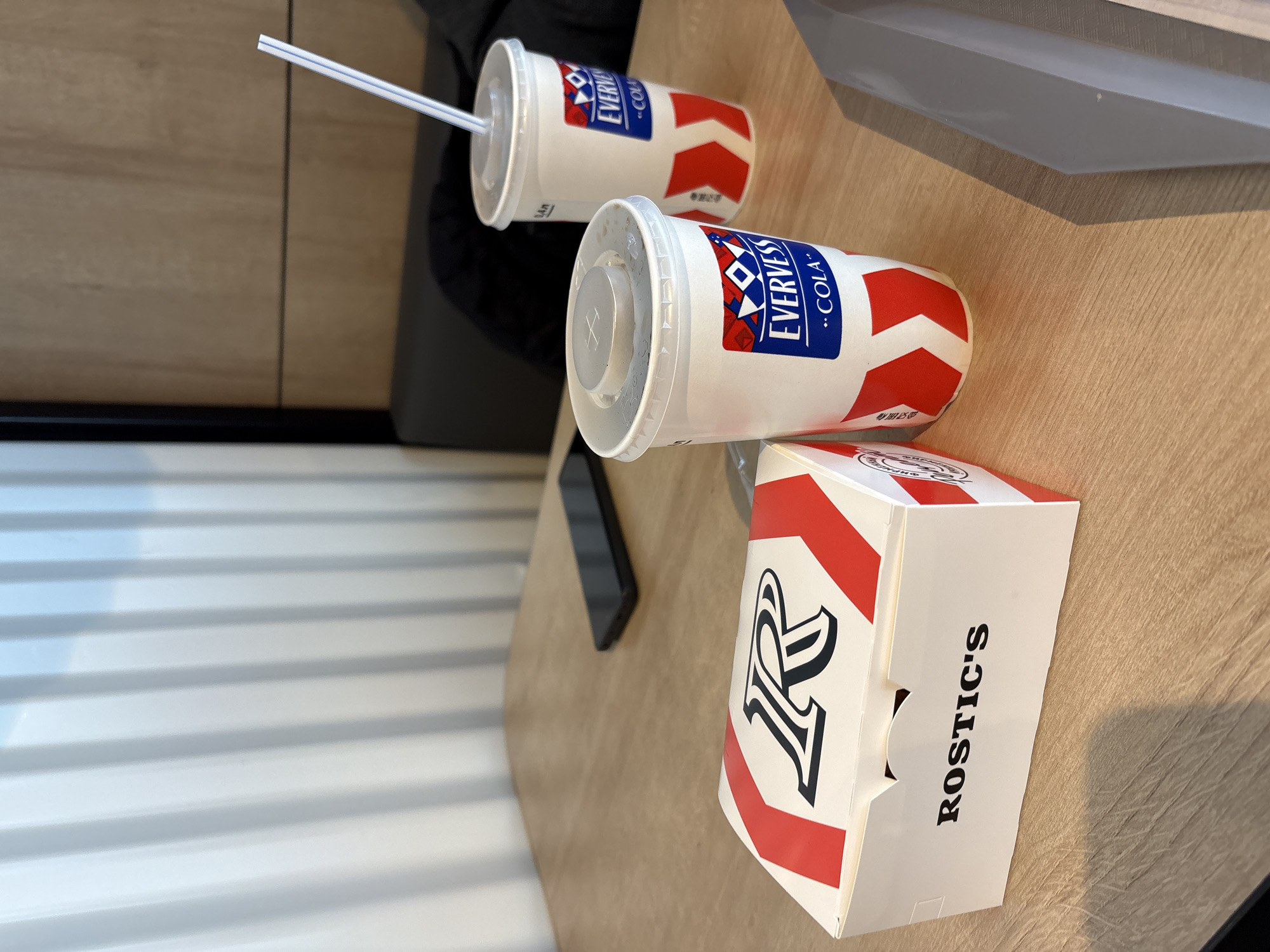 On a tan table sits a fast-food meal: two "Evervess" cola cups with lids, one with a straw, and a "Rostics'" branded food box. A black smartphone is also on the table. The scene likely takes place in a Rostics' restaurant in Krasnoyarsk, Russia. There are no people visible in the frame. The environment includes a corrugated wall.
