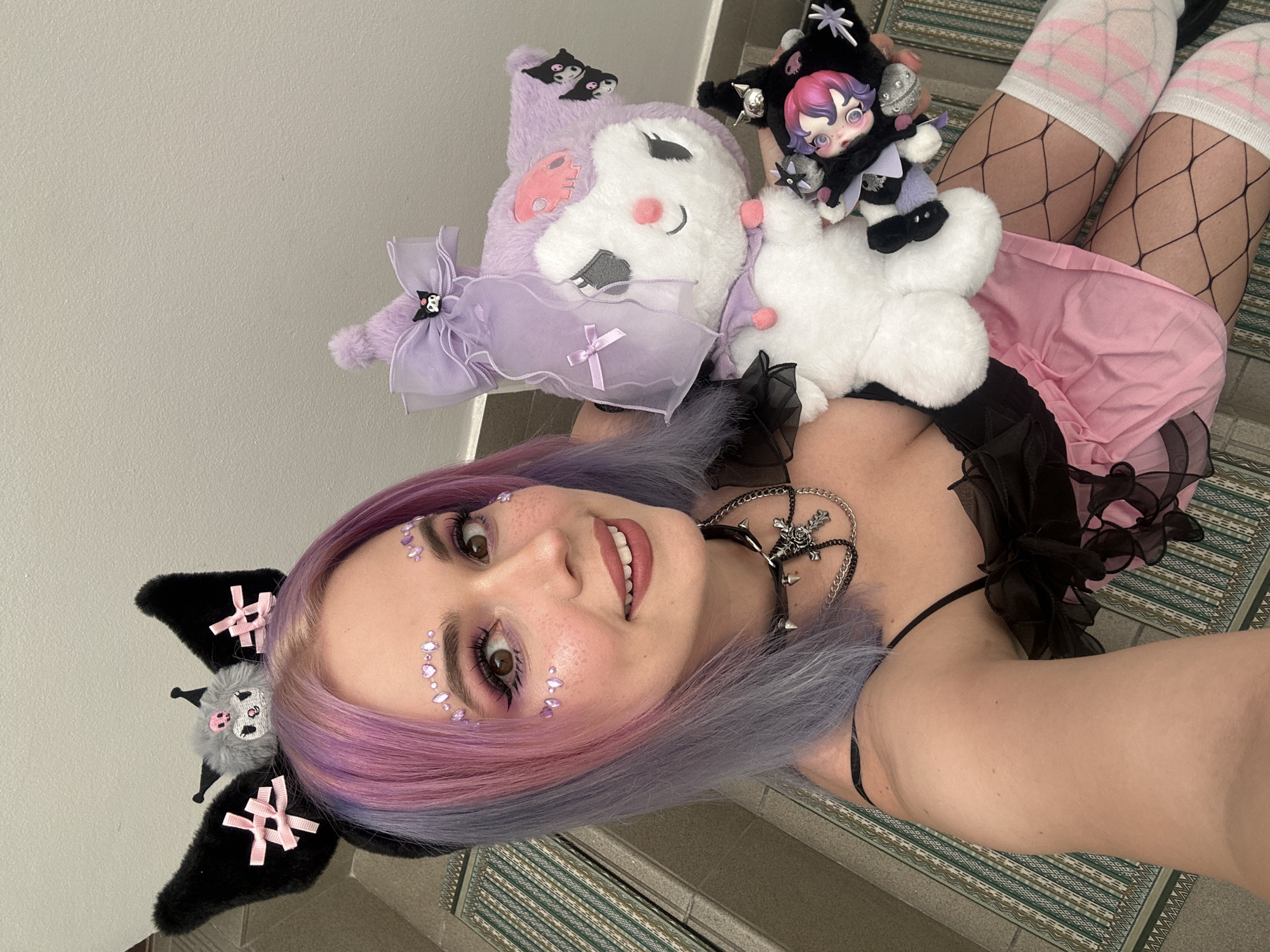 A person with lavender and pink hair, wearing a black choker, a black top, a pink skirt, fishnet stockings, and white striped socks, is holding three stuffed toys. The stuffed toys are purple, white, and black. The person has purple gems as makeup around their eyes. They are wearing a black headband with bat-like ears that have pink bows on them. The background appears to be a tiled wall. It's unknown if this image was taken in City of Zagreb, Croatia, as there are no discernible landmarks or features to indicate the location.