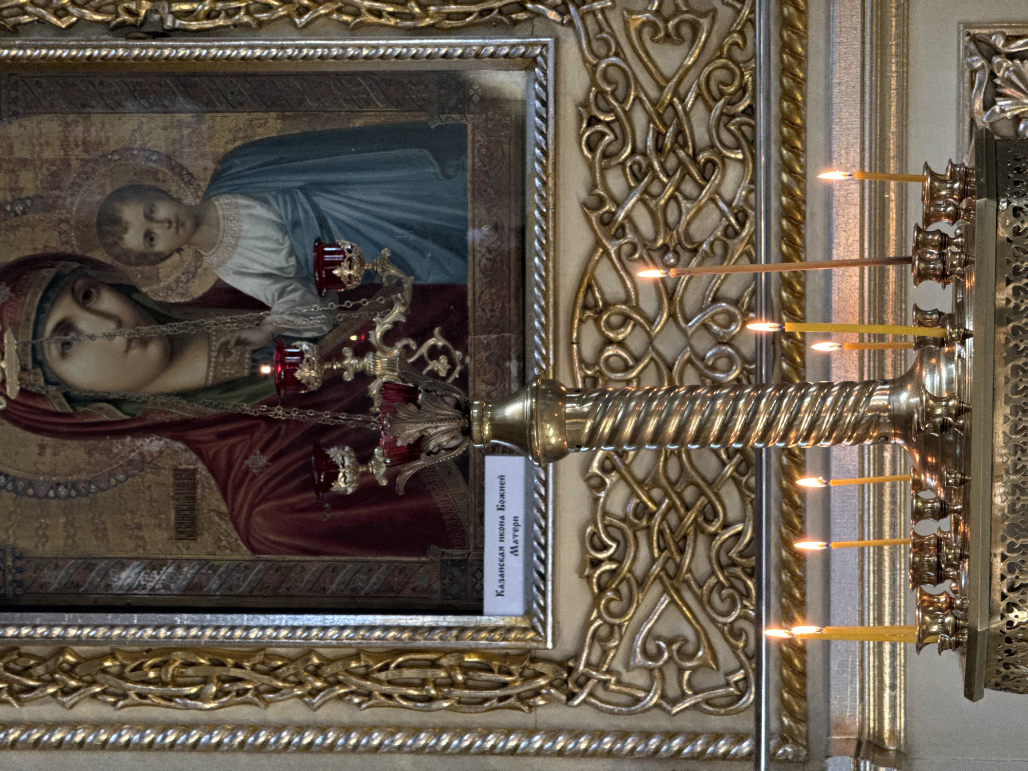 An Orthodox icon of the Virgin Mary and Christ Child is displayed within an ornate gold frame. Below the icon, a label reads "Казанская икона Божией Матери" (Kazan Icon of the Mother of God). In front of the icon, a brass candelabrum holds several lit yellow candles. The candelabrum is intricately decorated with twisting metalwork. The scene is set in what appears to be a church or religious sanctuary.