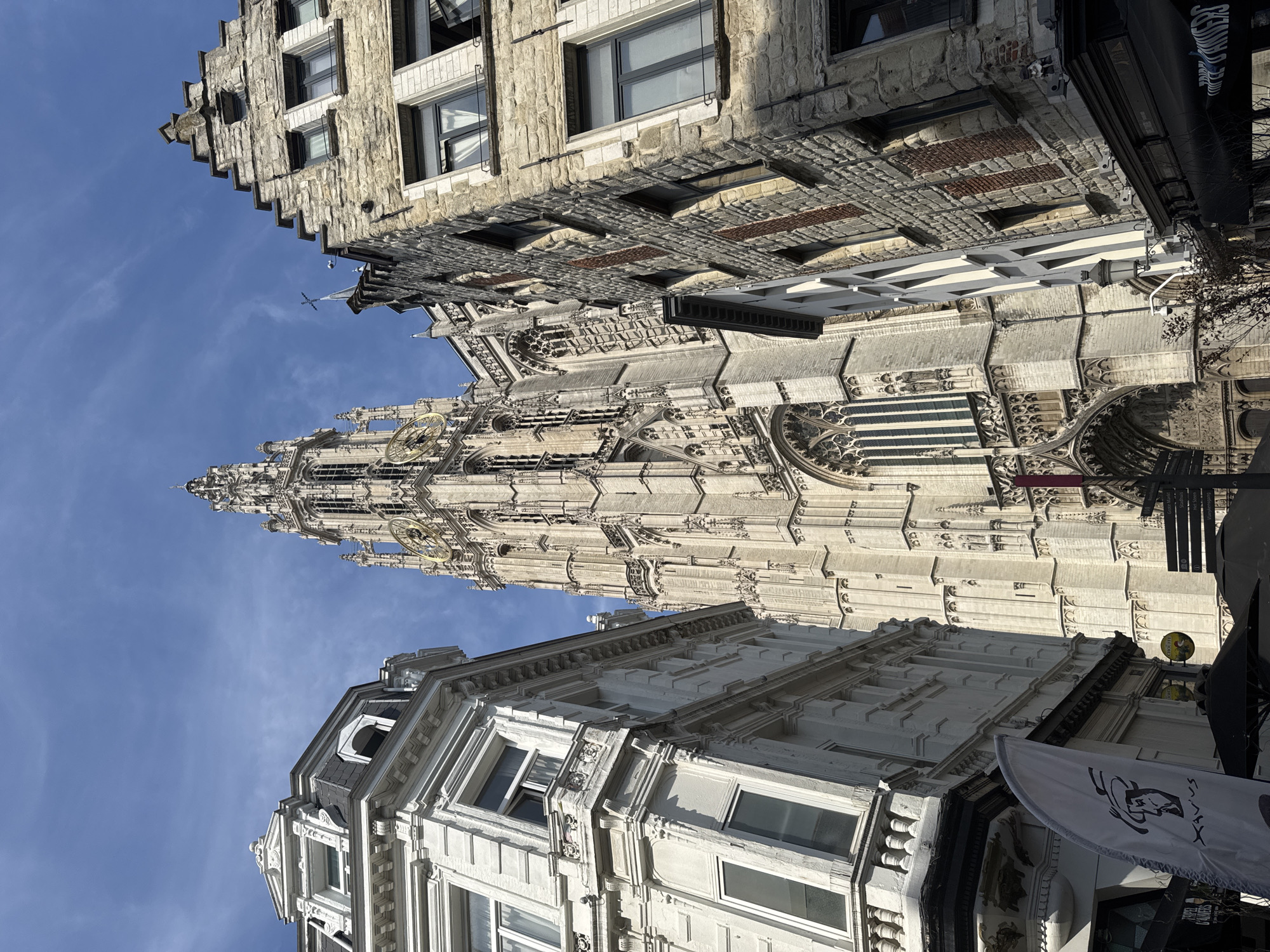 This is a low-angle, outdoor, daytime shot of the Cathedral of Our Lady in Antwerp, Belgium, as seen from a narrow street. The Gothic cathedral, made of pale stone, dominates the center of the frame with its tall, ornate spire. To the left is a white, ornate building with multiple windows. To the right is a building constructed of rough, grey stone with a stepped gable roof and red brick accents. The sky is blue with scattered white clouds. There are no visible people in the frame.