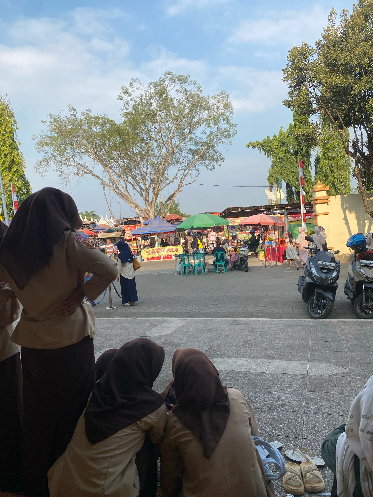 A crowd of people, some wearing hijabs and light brown uniforms, are gathered at an outdoor market in Nyamok, Indonesia. Several stalls with colorful awnings are set up, displaying various goods. Scooters are parked in the foreground. The sky is bright blue with scattered clouds, and trees with lush green foliage are visible in the background. Indonesian flags are displayed on poles. Some individuals are seated on the ground, facing away from the camera, while others stand and interact with vendors. The ground is paved with stone tiles.