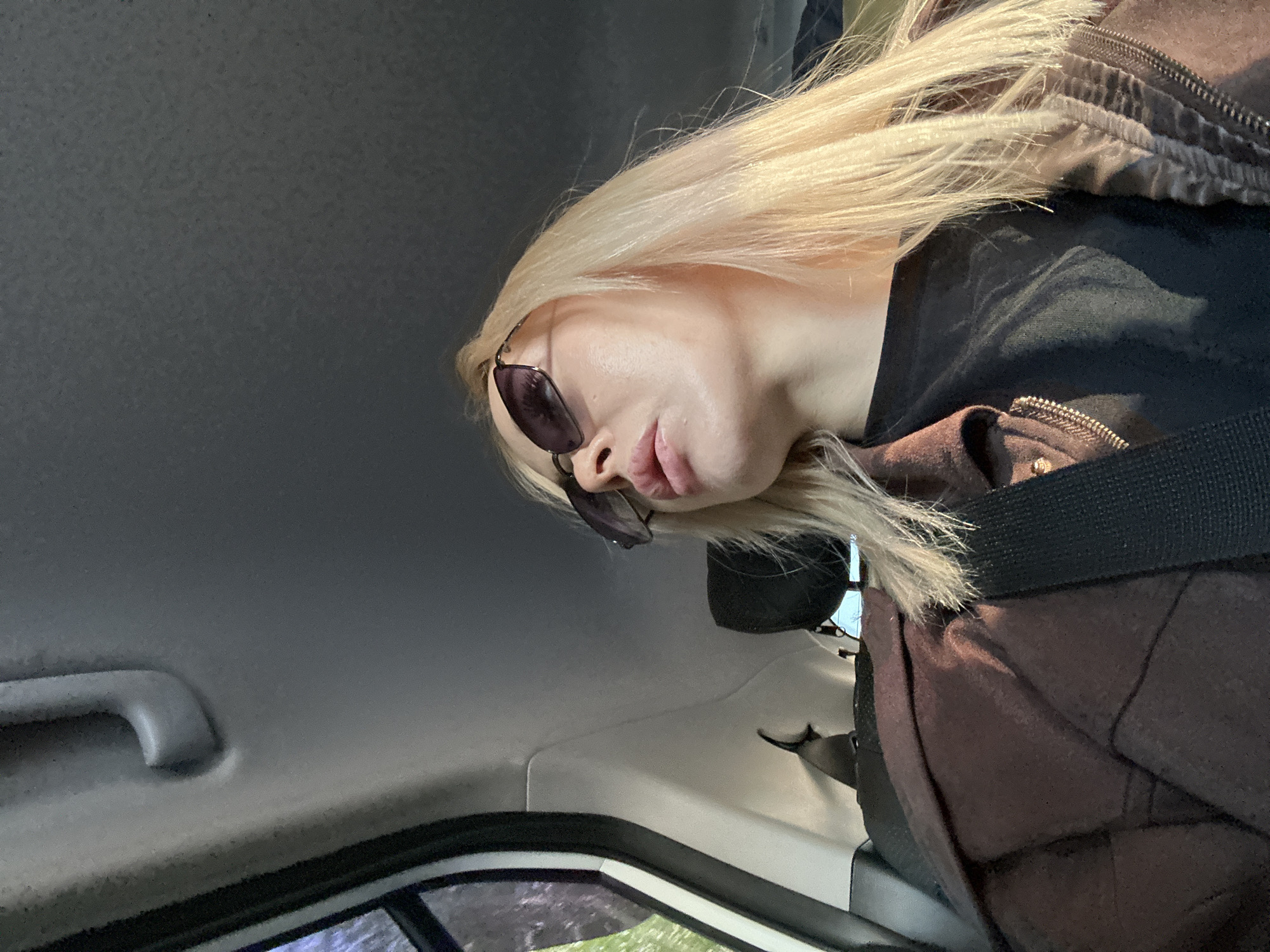 A woman with blonde hair and sunglasses is seated inside a vehicle. She is wearing a brown jacket over a black shirt and a black seatbelt. The interior of the vehicle is visible, including a gray ceiling and a window showing green vegetation outside. The image was taken from an angle, showing the woman's face and upper body. The location of the image is not discernible.