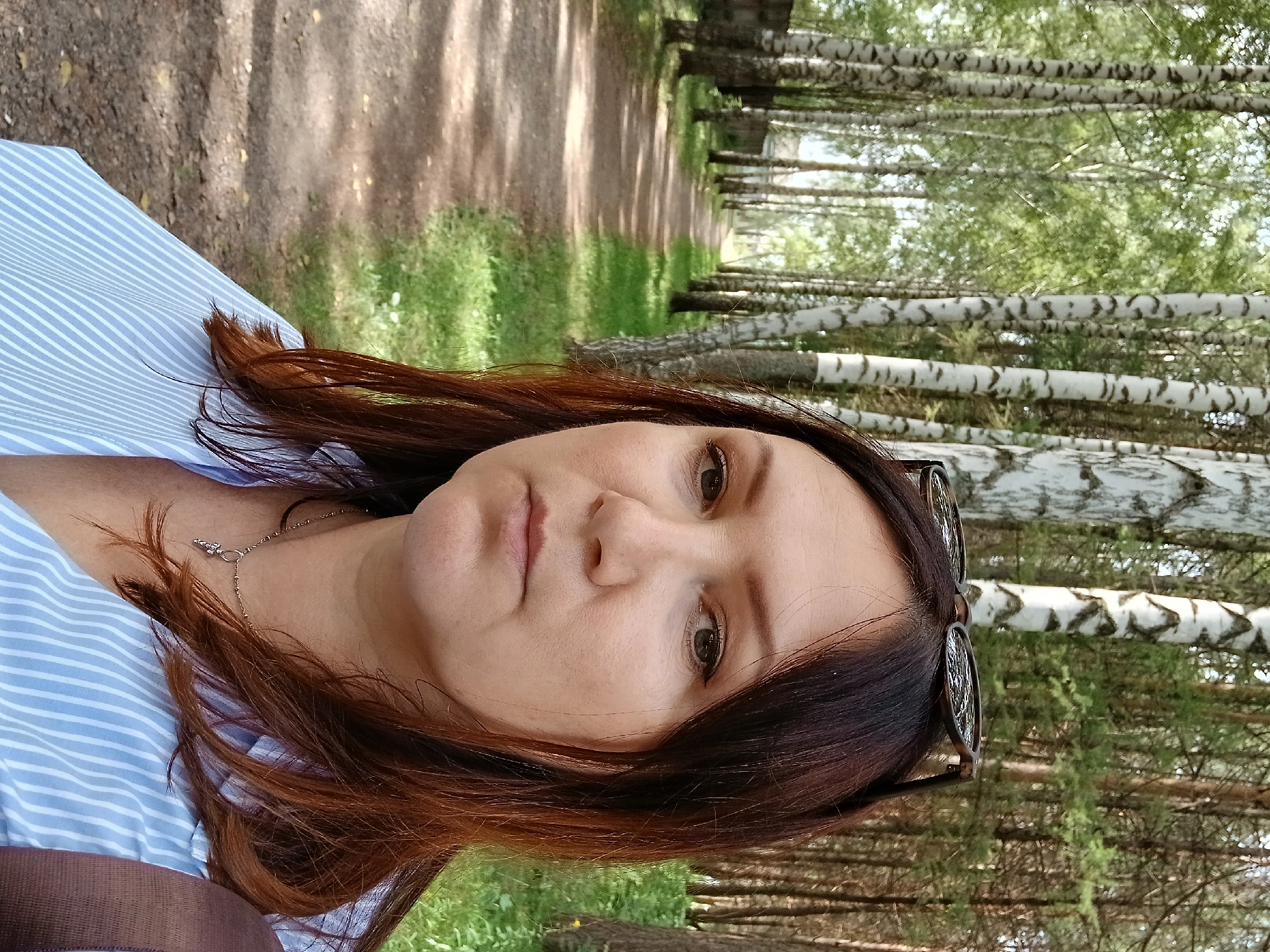 A woman with long, auburn hair and light brown eyes is taking a selfie outdoors. She is wearing a light blue and white striped button-down shirt and a silver necklace with a charm. Sunglasses are perched on top of her head.
The woman is standing on a dirt path lined with trees. On the left side of the path, there are birch trees with white bark and green leaves, and a low concrete wall. On the right side, there are pine trees with green needles. The path appears to be in a park or a wooded area within the city of Osa, Russia. The lighting suggests it is daytime, possibly late morning or early afternoon. The background shows more trees and foliage, indicating a natural environment. The overall scene is peaceful and outdoorsy.