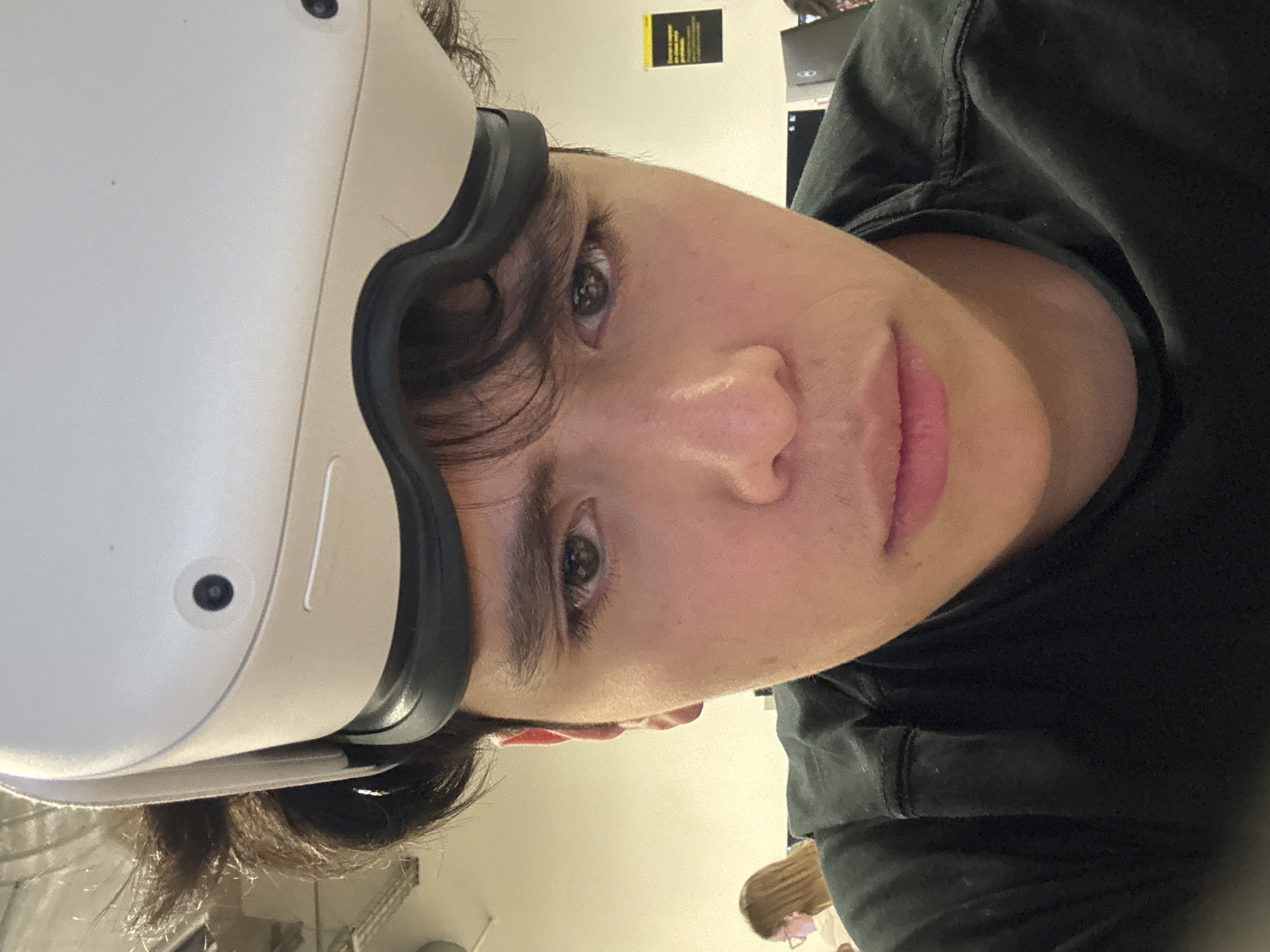 A close-up, angled selfie shows a person wearing a white virtual reality headset with a black trim. The person has fair skin, brown eyes, and dark hair. They are wearing a black shirt. The background is blurry, but a wall with a yellow sign and some office equipment are visible. Another person is partially visible in the background. The location is Mexico City, Mexico.