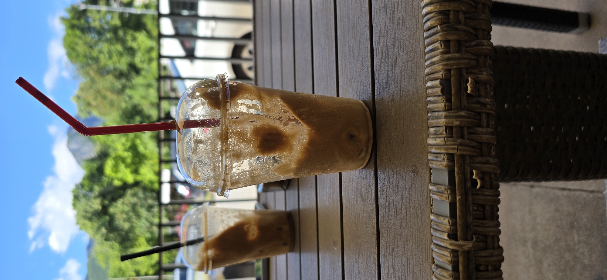 This is a vertical, close-up, outdoor photograph taken from a low angle, showing two plastic cups of iced coffee on a wooden table. The cup in the foreground is larger and slightly to the right, featuring a red straw extending upwards. The cup in the background is smaller and to the left, with a black straw. Both cups are partially filled with a brown, creamy liquid, and condensation is visible on the sides. The table is made of dark brown wooden planks. Behind the table, a metal railing is visible, and beyond that, lush green trees, a mountain, and a blue sky with white clouds. In the background, the blurry shapes of white vehicles suggest a parking lot or street. The foreground of the table has a woven wicker edge.