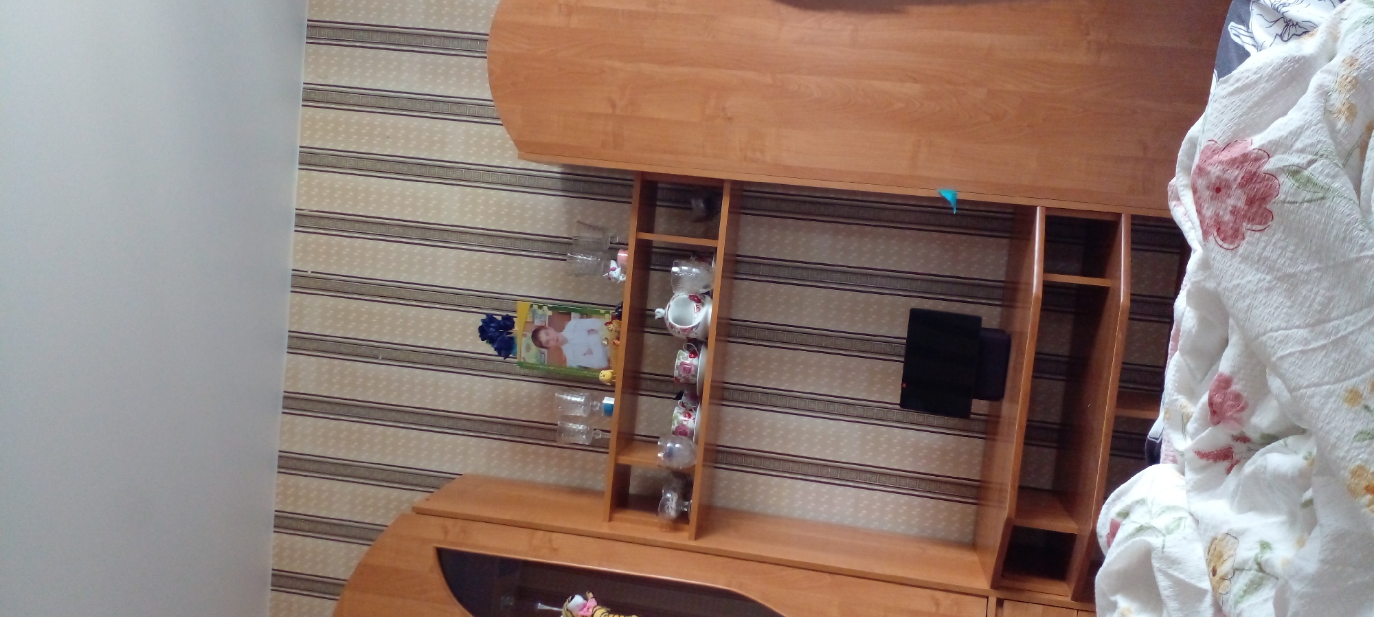 The image shows an interior scene, likely a bedroom or living room, featuring a wooden cabinet with shelves. On the shelves are various items including glasses, cups, saucers, a teapot, a framed picture of a young boy, and a small black electronic device. The walls are covered with striped wallpaper. In the foreground, a white blanket with floral patterns is visible. The context of Glubokoe, Kazakhstan, is not visually ascertainable from the image itself.