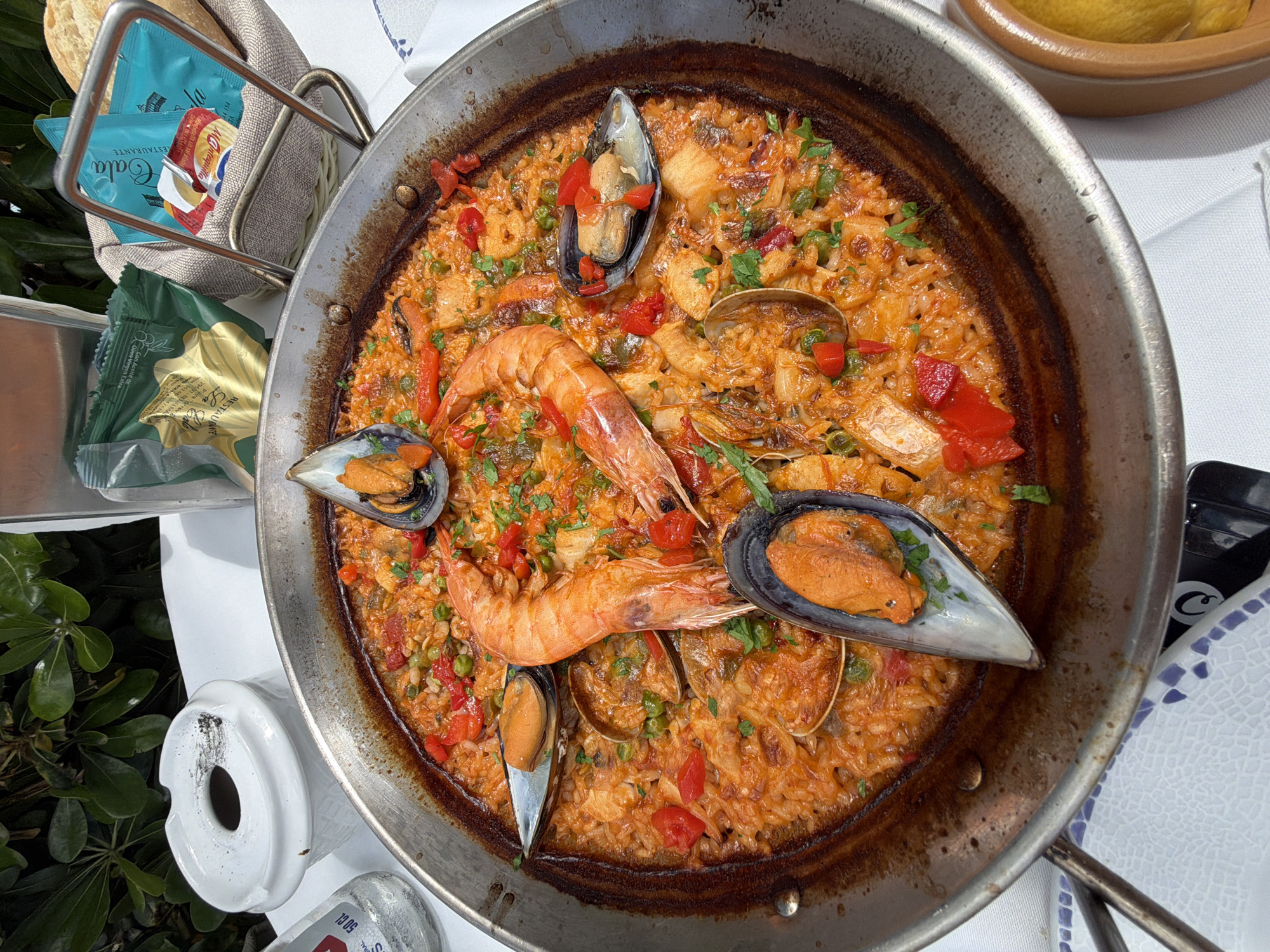 The image shows a close-up, overhead view of a paella in a large metal pan, likely served outdoors. The paella contains rice, mussels, shrimp, clams, red peppers, and parsley. The pan has a burnt edge and sits on a white-covered table. To the left of the paella, there is a small basket with bread and butter packets, and a white porcelain object. In the lower left, there's a glimpse of green foliage. The butter packets are labeled "RESTAURANTE Cala," which indicates the image was taken at a restaurant named Cala, referencing Playa Torrequebrada, 29630-BENALMÁDENA, in the city of Benalmádena, Spain.