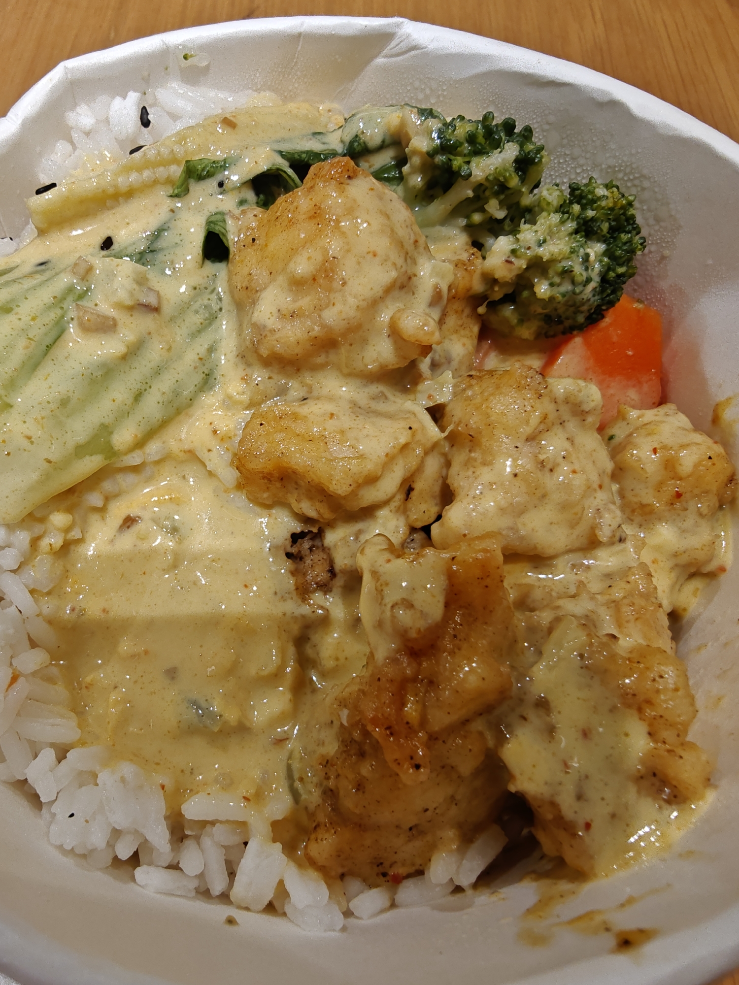 A close-up overhead shot shows a bowl of rice topped with a creamy yellow curry sauce, pieces of golden-brown fried chicken, broccoli florets, and what appears to be baby corn and spinach. The food is presented in a white paper bowl. The lighting is warm and diffused, highlighting the textures and colors of the meal. The background is out of focus, suggesting a casual dining setting, possibly in a restaurant or food stall. The image is static, with no people or actions depicted. This image does not provide any information about its location in Dubai, United Arab Emirates.