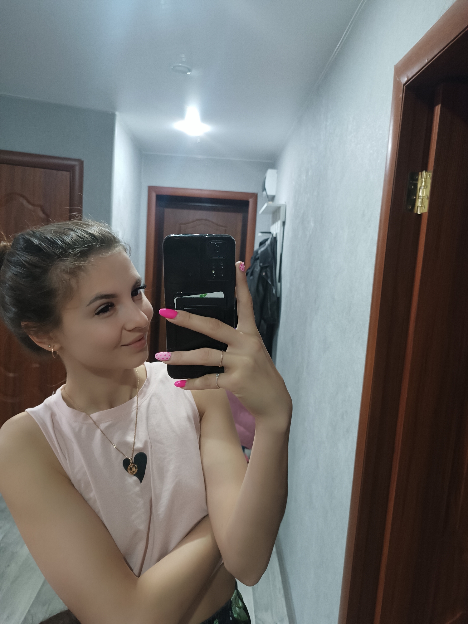 A young woman is taking a selfie in a hallway of a residence, location unknown. She is holding a black smartphone with a cardholder attached to the back, her right hand is visible with pink nails, one with a polka-dot design. She is wearing a light pink sleeveless top with a heart-shaped pendant necklace. Her hair is pulled back in a bun. The hallway has two closed wooden doors and a light fixture on the ceiling. A white wall with shelving and hanging clothes is visible behind her in the reflection of a mirror.