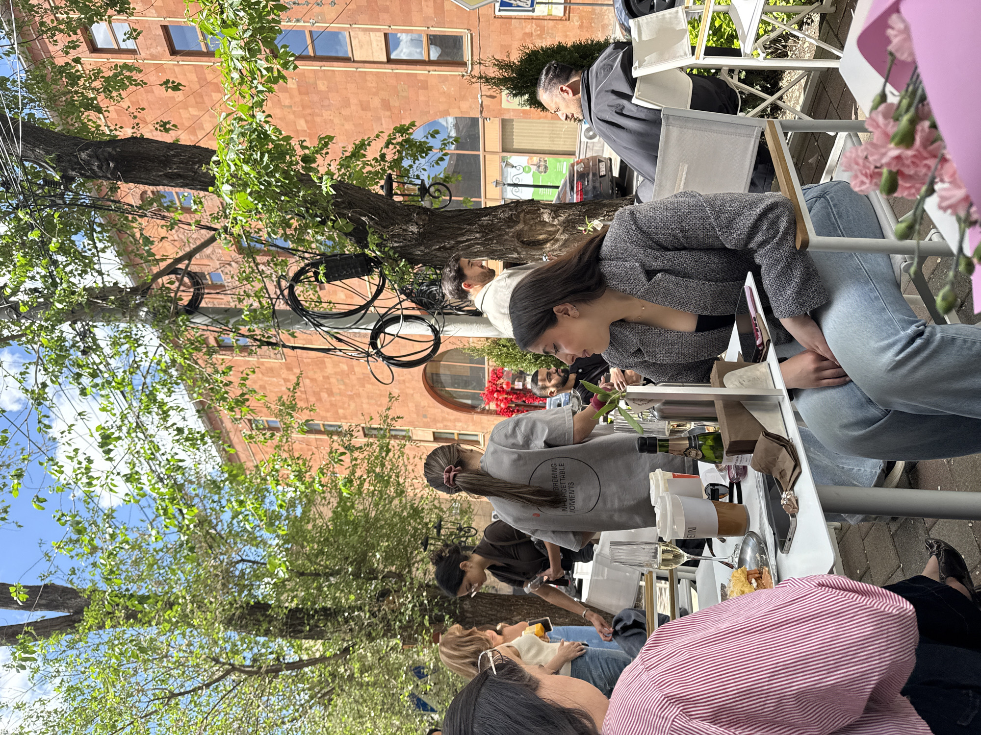 The image captures an outdoor dining scene, likely at a café or restaurant, during daytime. The location is recognizable as Yerevan, Armenia, suggested by the architectural style of the brick building in the background. Several people are seated around tables, with some visible in conversation and interacting. One person is wearing a grey t-shirt, and another is wearing a grey blazer. The tables are covered with items typically found in a restaurant setting, including wine bottles, glasses, food, napkins, and other tableware. There is a tree with green foliage providing shade over the area, with electrical wires running through its branches. A bouquet of flowers sits on a table.