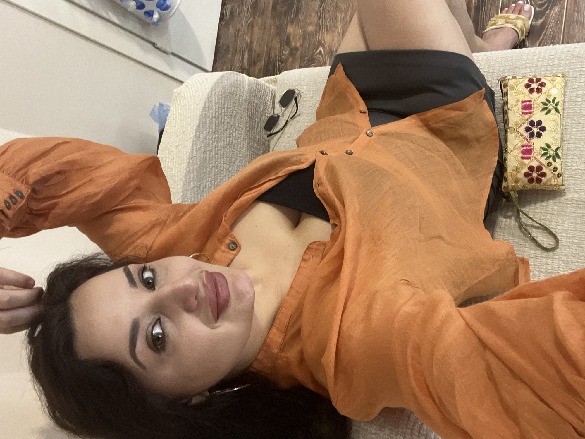 In an indoor setting, a woman with dark hair and olive skin is captured from a high angle, reclining on a couch. She is wearing an open, loose-fitting orange button-down shirt over a black top, revealing her décolletage. Her lips are painted with a dark pink lipstick, and she wears hoop earrings. A small, embroidered clutch with floral patterns rests on the couch beside her, along with a pair of sunglasses. Her right hand is raised, with fingers touching her hair, and her left arm is extended. The background shows a wooden floor and a white wall. The woman's legs are visible, and she is wearing gold embellished sandals.