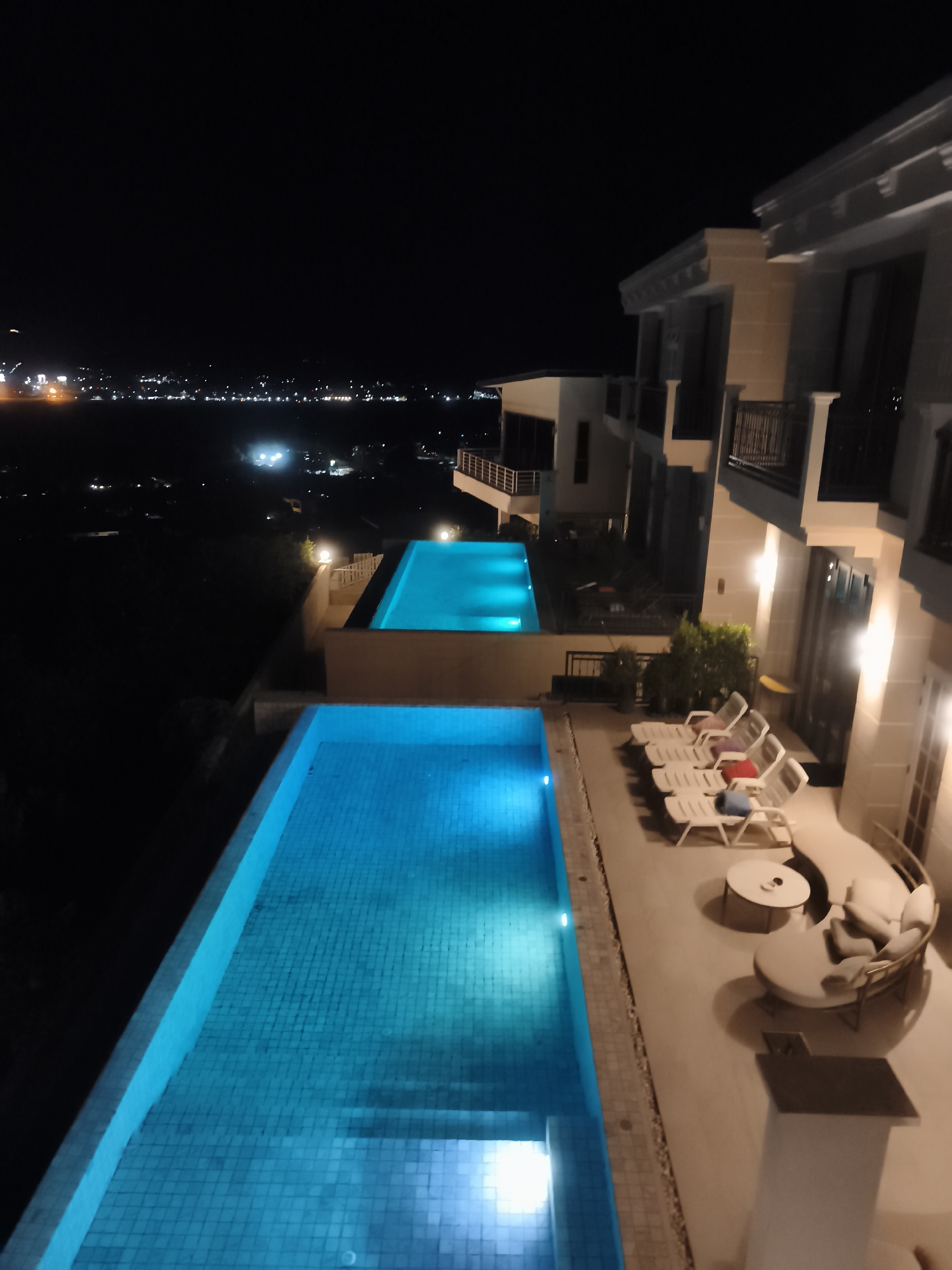 This is a night time aerial view of an outdoor infinity pool area at a resort in Baan Bang Rak, Thailand. The pool is illuminated with a vibrant turquoise glow from underwater lights, reflecting on the tiled bottom. The scene is framed by a modern, multi-story building with balconies and outdoor lighting. In the foreground, several white lounge chairs are neatly arranged on a patio, some with colorful cushions. A circular coffee table sits in front of a large, curved outdoor sofa with plush cushions, suggesting a relaxing atmosphere. Beyond the pool, a distant city skyline is visible, dotted with lights, indicating an urban or developed area in the background. The overall impression is one of luxury and tranquility, suitable for a vacation or a peaceful evening retreat. The time of day is clearly night, with the darkness contrasting with the illuminated pool and building lights.