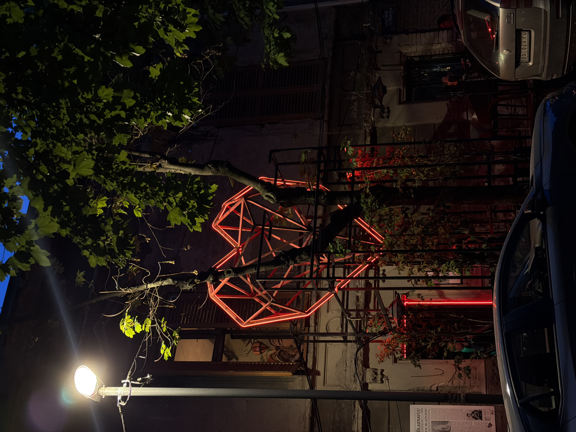 A street scene at night in Belgrade, Serbia. An illuminated, geometric red heart sculpture is affixed to a tree branch in front of a building. A streetlamp provides bright illumination to the tree's lower branches. Two parked cars are visible on the right side of the frame. A sign with text and a black and white picture is visible at the bottom right. A building facade with plants and some red lighting is visible behind the tree.