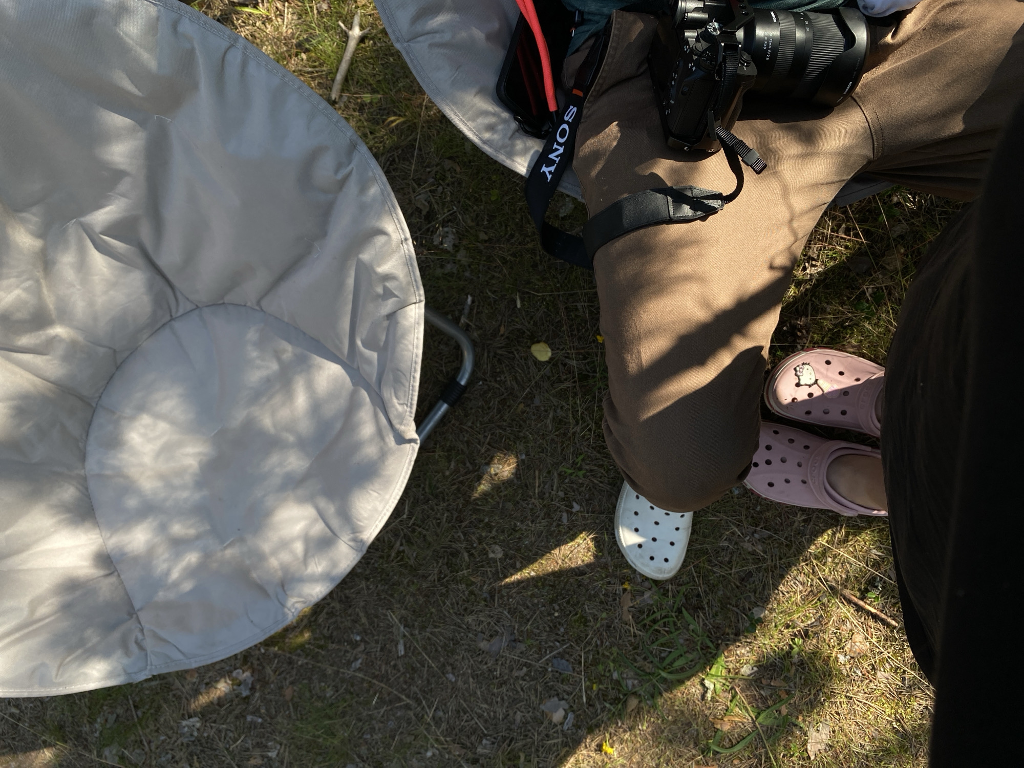 The image depicts a ground-level perspective of a person's legs and feet, positioned outdoors in what appears to be a natural setting. The individual is wearing brown trousers, pink Crocs with a Hello Kitty charm on the left shoe, and a white Croc on the right foot. A black camera with a lens is held in the right hand, and a black Sony camera strap is visible. The background includes dry grass, dirt, and two light-colored outdoor chairs. Sunlight casts shadows across the scene. No specific identifiable landmarks or features place this scene within the city of Primorsk, Russia.