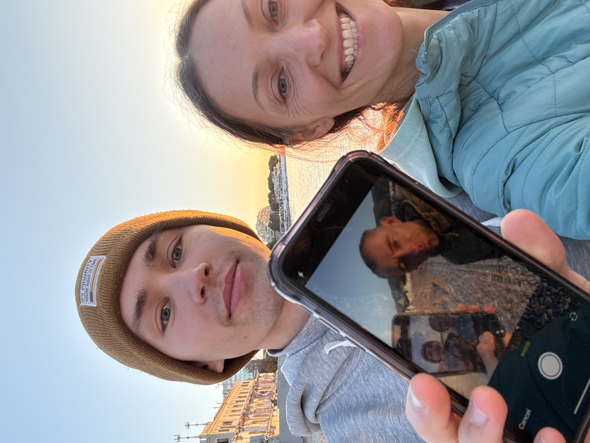 The image is a rotated outdoor selfie featuring a woman and a man, captured during golden hour or sunset with a clear sky.
The woman, positioned in the upper right quadrant of the frame, smiles broadly, revealing her teeth. She has dark hair and wears a light blue puffer jacket over a lighter blue layer. Her face is illuminated by warm, soft light.
The man, in the lower left, looks directly at the camera with a neutral expression. He wears a brown knit beanie with a white label that reads "IT'S PRIORITY YOU'RE RIGHT IN TIME". He also wears a grey hooded sweatshirt.
He holds a black smartphone prominently in the center foreground. The phone's screen displays a clear reflection of the man, the woman, and the background scene, including buildings along a waterfront and the sun-kissed water. The reflection creates a complex, recursive image. The phone's camera interface is visible on the screen, showing "PHOTO" and "Cancel" buttons.
The background reveals a waterfront setting, with several buildings lining the shore of a body of water, likely a bay or sea. The sky transitions from light blue at the top to warmer orange and yellow tones near the horizon, indicating the time of day. The location is Gelendzhik, Russia.