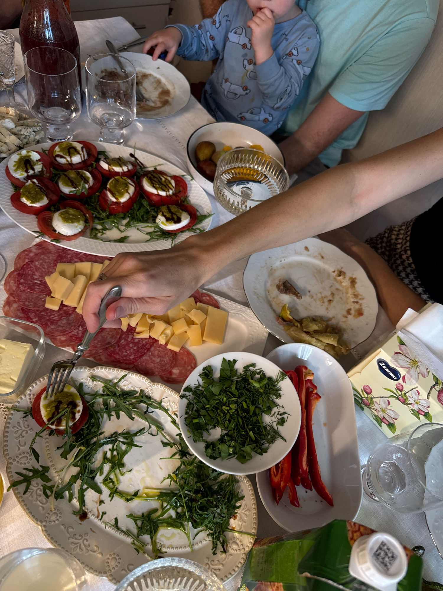 The image depicts a close-up, overhead view of a table laden with food and drinks, suggestive of a meal in progress. The setting is likely domestic, with a white tablecloth as the primary surface. On the table are plates of caprese salad, salami, cubed cheese, sliced red bell peppers, and a bowl of chopped herbs. There are also several glasses, a bottle of dark liquid (possibly wine or juice), and a juice carton. In the background, a young child in a blue onesie with car prints is seated and reaching towards the table. An adult's arms are visible, one resting on the table, and another person's arm is reaching for food. The scene suggests a casual gathering or family meal. The location is noted as Захарьино, Russia, though this detail is external to the visual information provided by the image itself.
