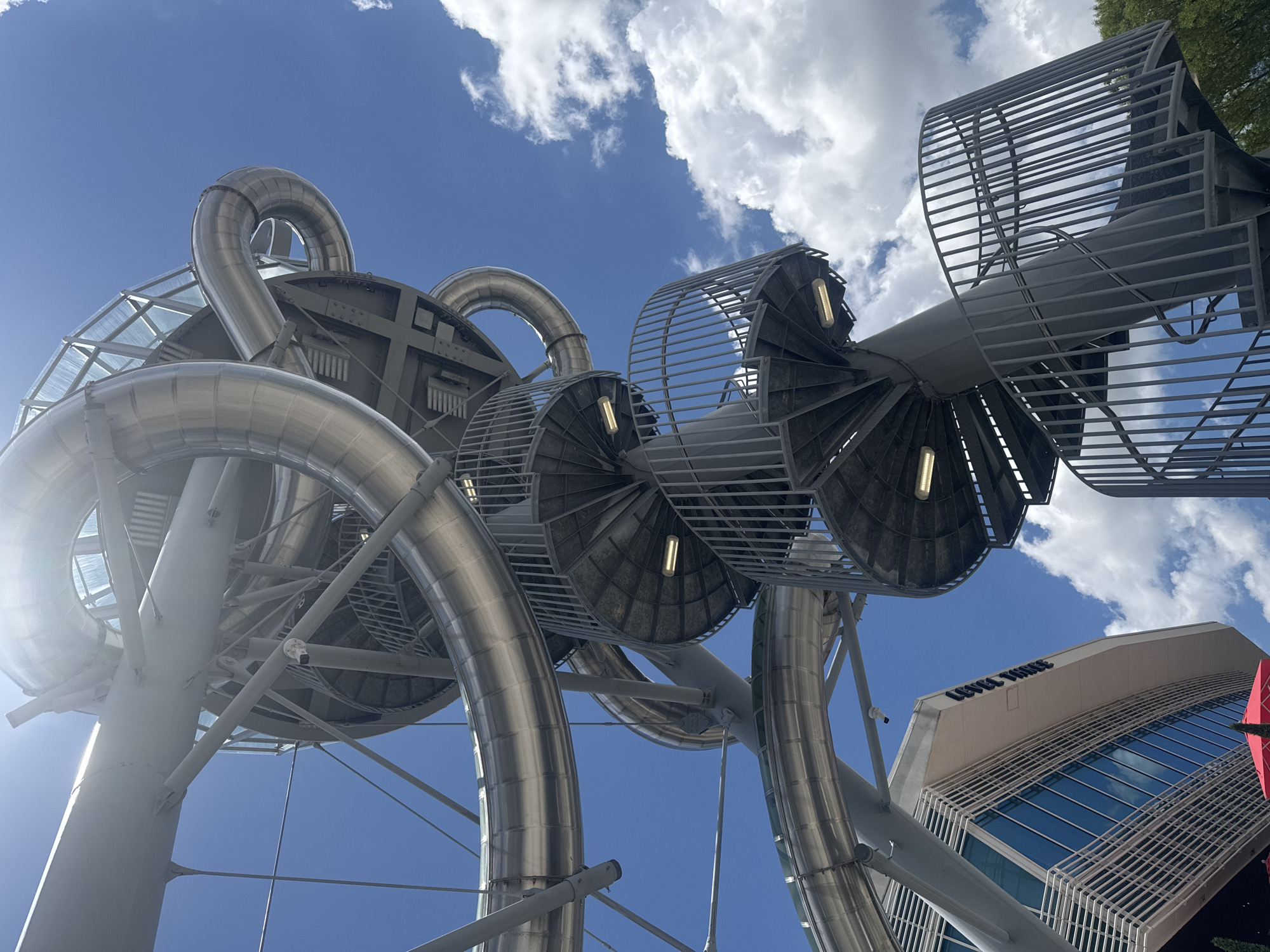 The image presents a low-angle view of a large, intricate metal structure with curved pipes and circular platforms. The structure appears to be an art installation or a unique architectural feature. Each circular platform has interior lighting. The structure is set against a backdrop of a blue sky with scattered white clouds. A building with the words "LEVEL THREE" on its side can be seen in the lower right corner of the image. The location is in the city of Aventura, in the United States of America.