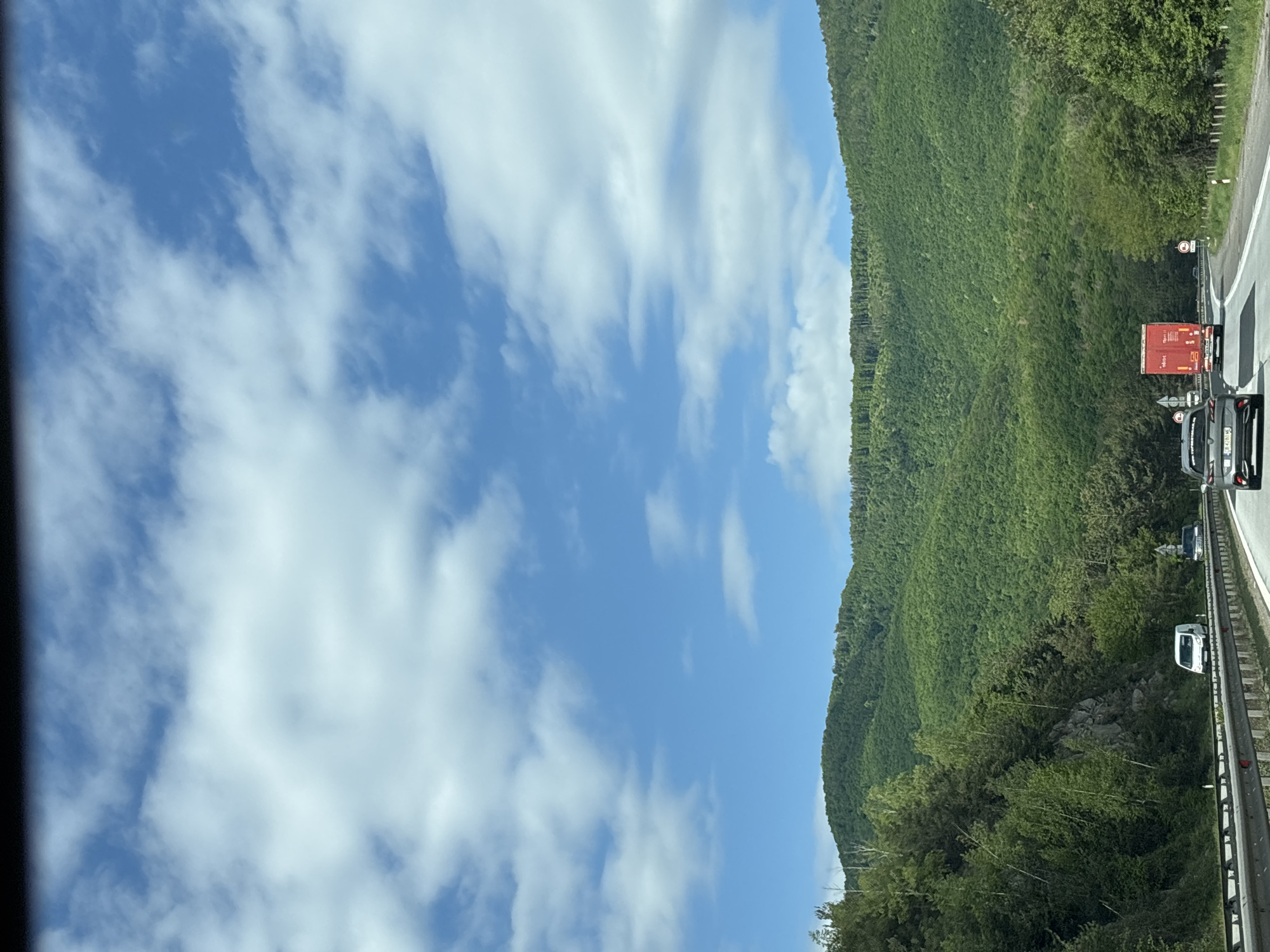 The image shows a highway scene taken near Dolno Varshilo, Bulgaria. A lush green mountain slopes upward on the right, contrasting with a partly cloudy blue sky on the left. A highway runs alongside the mountain, and a number of vehicles, including a red truck and a gray van, are on the highway. A metal guardrail separates the highway from the mountain side.