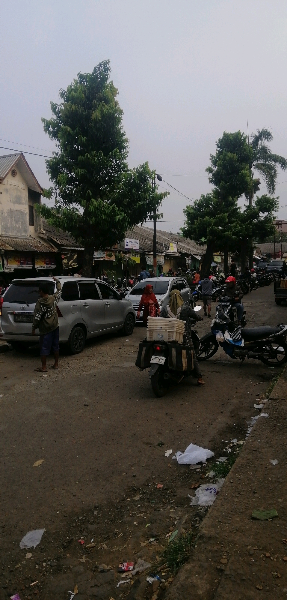 The image depicts a street scene in Ajibarang, Indonesia, characterized by a mix of vehicles, buildings, and vegetation under an overcast sky. In the foreground, a person in a green and white striped shirt and blue shorts stands near a silver car, with their right arm raised. A silver minivan is parked behind them. To the right, a motorcycle is stopped, carrying a white crate on its rear rack, with a person in a red hijab and another in a light brown hijab walking near it. Further down the street, a line of parked motorcycles is visible. Commercial buildings with corrugated metal roofs line the left side of the street, featuring various signs and advertisements. Several trees, including a tall palm tree, are scattered throughout the scene. The ground is a mix of dirt and pavement, with visible debris and trash. The overall atmosphere suggests a busy, perhaps market-like, environment in a moderately populated area.