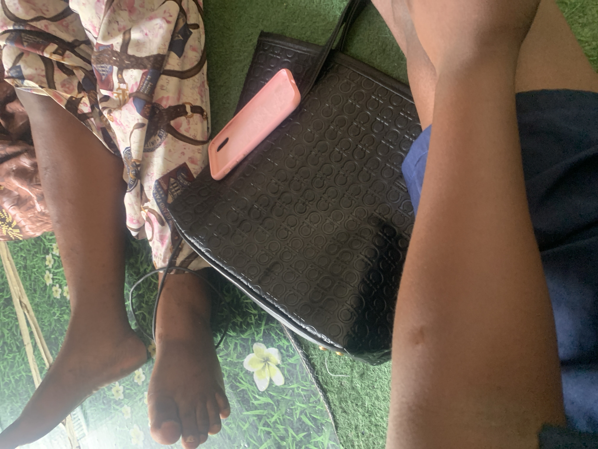The image depicts a close-up, low-angle view of two individuals' legs and feet resting on a green surface that resembles artificial grass. A black, textured handbag is positioned in the center of the frame, with a pink phone case resting on top of it. To the left of the handbag, a single white flower with yellow accents is visible on the green surface. In the background, the patterned fabric of clothing is visible, featuring a design of belts and floral elements. The scene appears to be an indoor or shaded outdoor setting, possibly in Jauro Yinu, Nigeria, based on the provided context. The lighting is moderate, creating soft shadows.