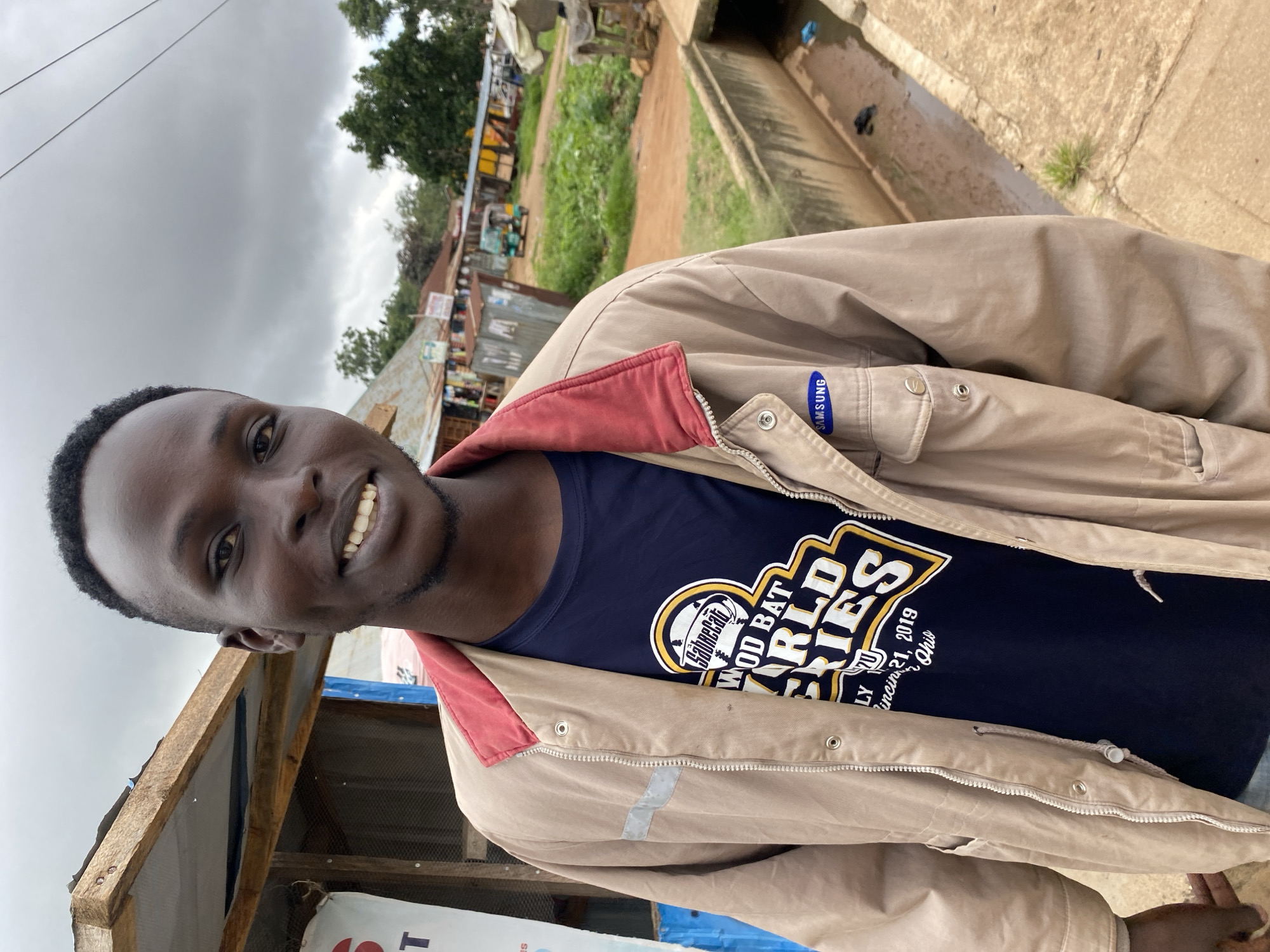 A young Black male with short hair and a light beard smiles at the camera. He is wearing a dark blue t-shirt with a yellow and white graphic that reads "WOOD BAT WORLD SERIES" and a tan jacket with a red collar. The jacket has a silver reflective stripe on the left chest and a blue Samsung logo on the right chest. He is standing outdoors in what appears to be a marketplace in Bukuru, Nigeria. In the background, there are various market stalls with goods displayed, some corrugated metal structures, and a dirt road. A small, green and yellow motorized rickshaw is visible in the middle ground. A concrete drainage channel runs along the right side of the frame. The sky is overcast.