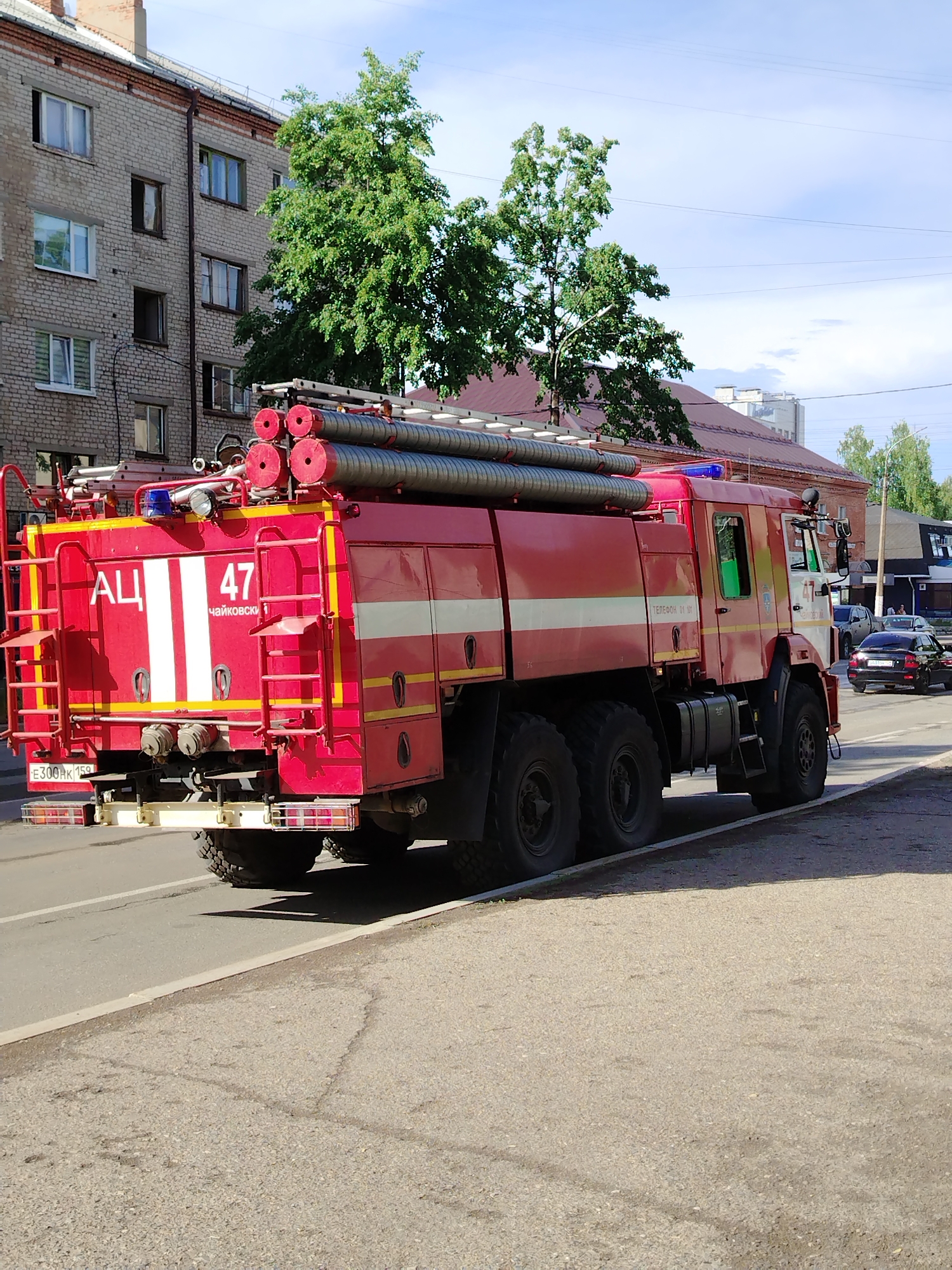 A large red fire truck, marked with "АЦ 47 Чайковский" and a license plate starting with "E300", is parked on the side of a street in Chaikovsky, Russia. The truck has numerous hoses and a ladder rack on top. A multi-story brick building with several windows is visible in the background, along with green trees. A dark-colored car is driving on the street behind the truck. The day appears sunny. The truck is parked along a curb next to a paved surface.