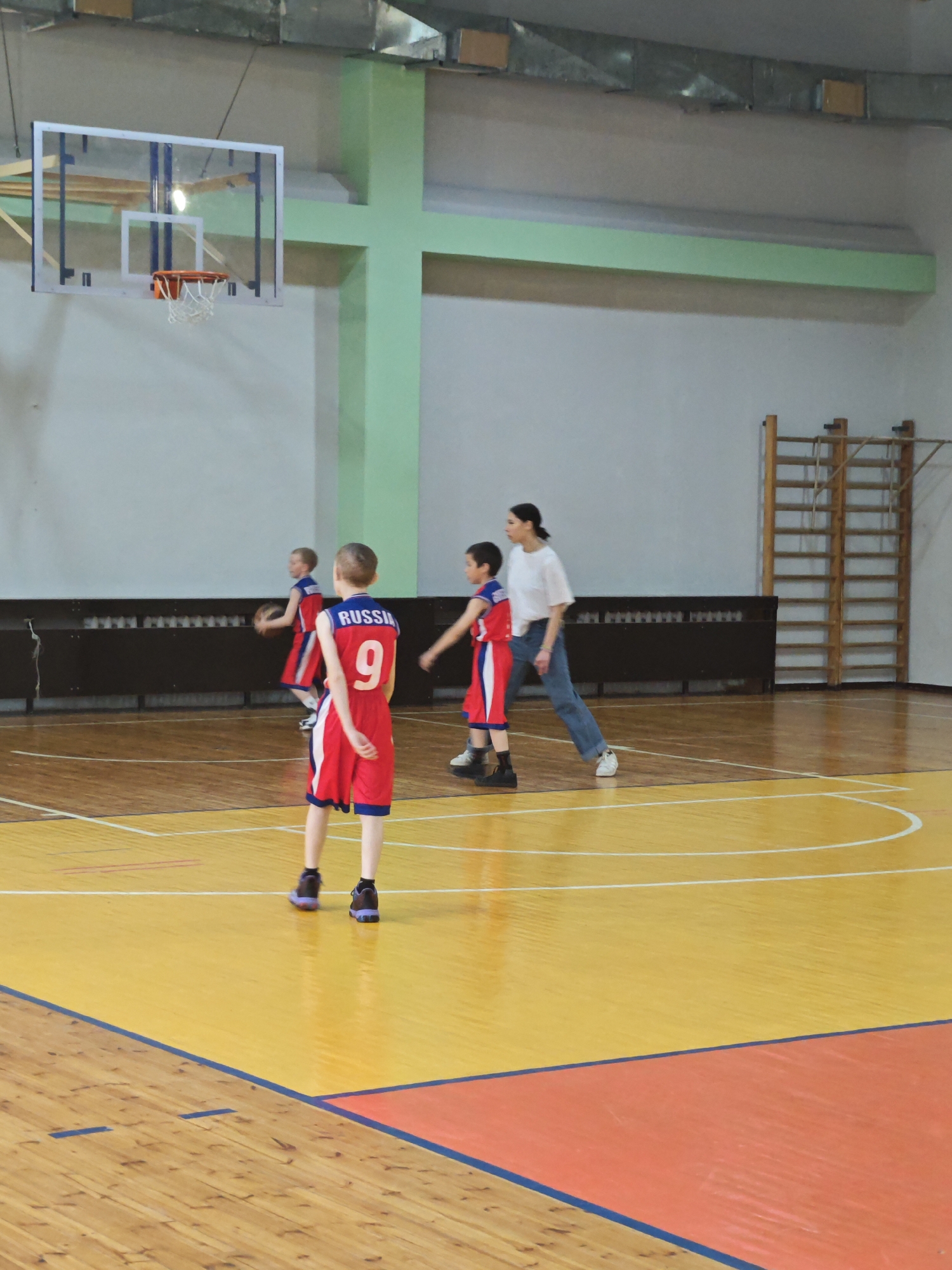 The image depicts a basketball court in what appears to be a gymnasium. Three young boys wearing red and white basketball uniforms with "RUSSIA" printed on the back are present, one of whom is wearing the number "9." A woman stands near them, dressed in a white shirt and blue jeans. The court's floor is varnished wood with distinct lines. A basketball hoop and backboard are visible. A set of wooden climbing bars is against one wall. The location is Markovskii, Russia.