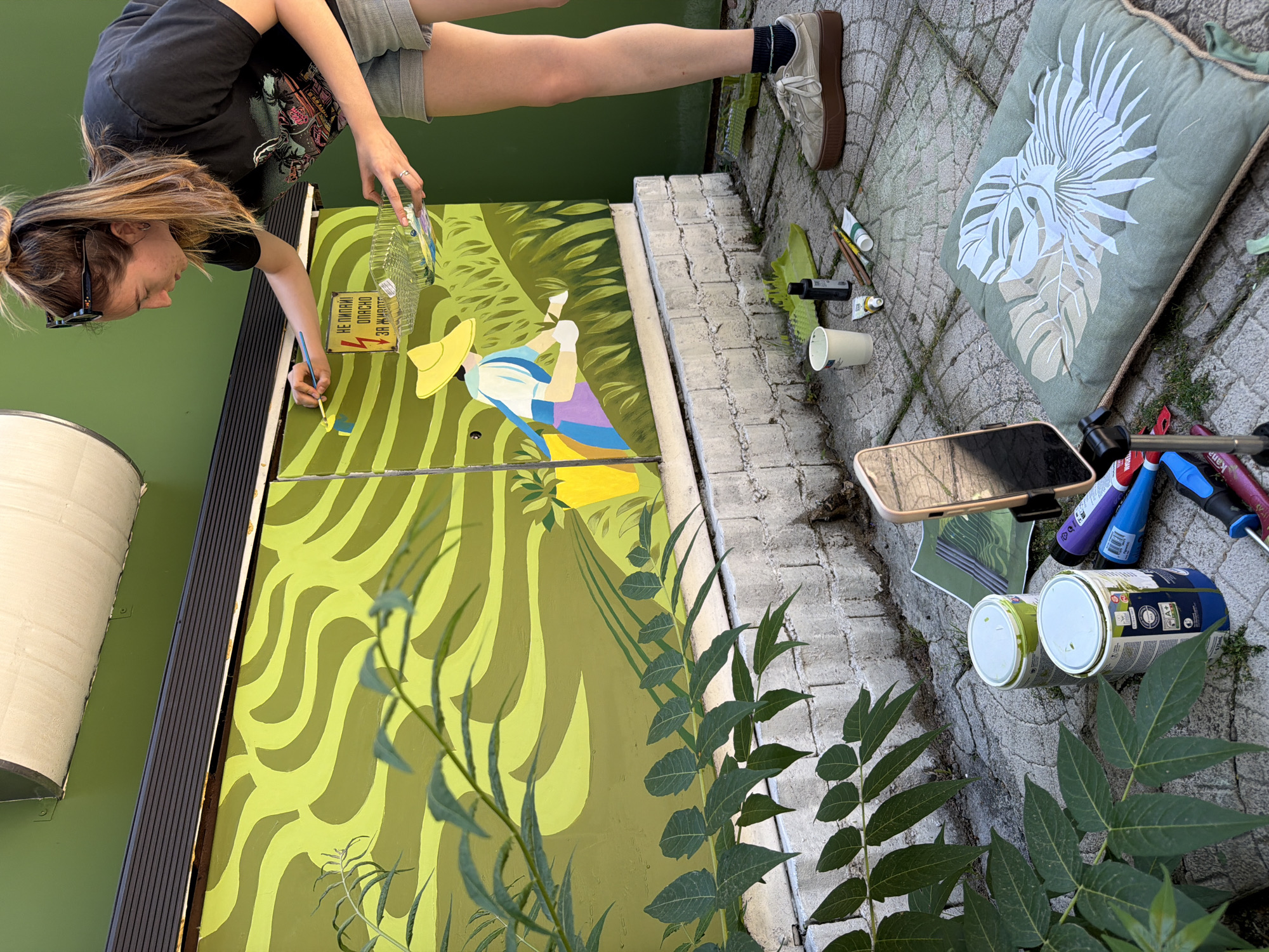 A young woman is painting a mural on a green, wavy background with shades of lime and olive green. She is wearing a black t-shirt and grey shorts, with sunglasses on her head. She holds a paintbrush in her right hand and a clear plastic container with paint tubes in her left. A yellow straw hat and a figure in blue and purple clothing are depicted in the mural. A yellow warning sign with Cyrillic text is also part of the mural.
In the foreground, various art supplies are scattered on a cobblestone path, including paint cans, brushes, tubes of paint, and a smartphone. A large, green cushion with a white leaf pattern rests on the ground. Green foliage is visible in the lower right and left corners of the image. The scene appears to be outdoors, possibly in a courtyard or alleyway, given the cobblestone and brick textures. The lighting suggests it is daytime, with no indication of weather conditions. The overall mood is one of creative activity.
Visible text on the warning sign reads: "НЕ ПИПАЙ! ОПАСНО ЗА ЖИВОТА!" which translates to "DO NOT TOUCH! DANGEROUS FOR LIFE!". Some text is also visible on the paint cans.