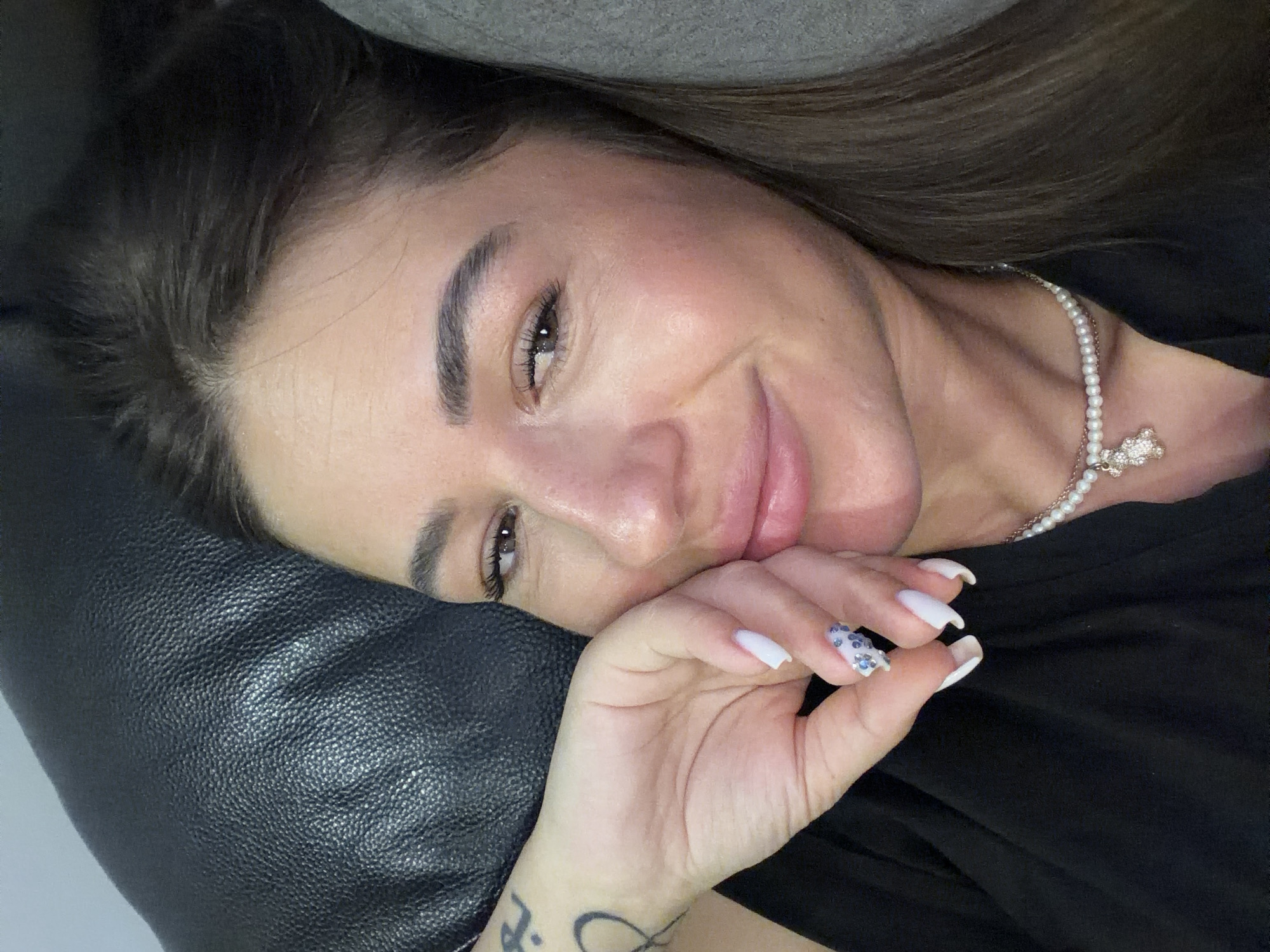 A woman with dark hair, wearing a black top and a pearl necklace with a teddy bear pendant, rests her head on a dark, textured surface, possibly a sofa. Her right hand is raised to her face, with manicured nails, one decorated with small blue stones. A tattoo is visible on her left wrist. She is looking towards the camera with a slight smile. The scene appears to be indoors. There is no indication of the city of Novi Iskar, Bulgaria.